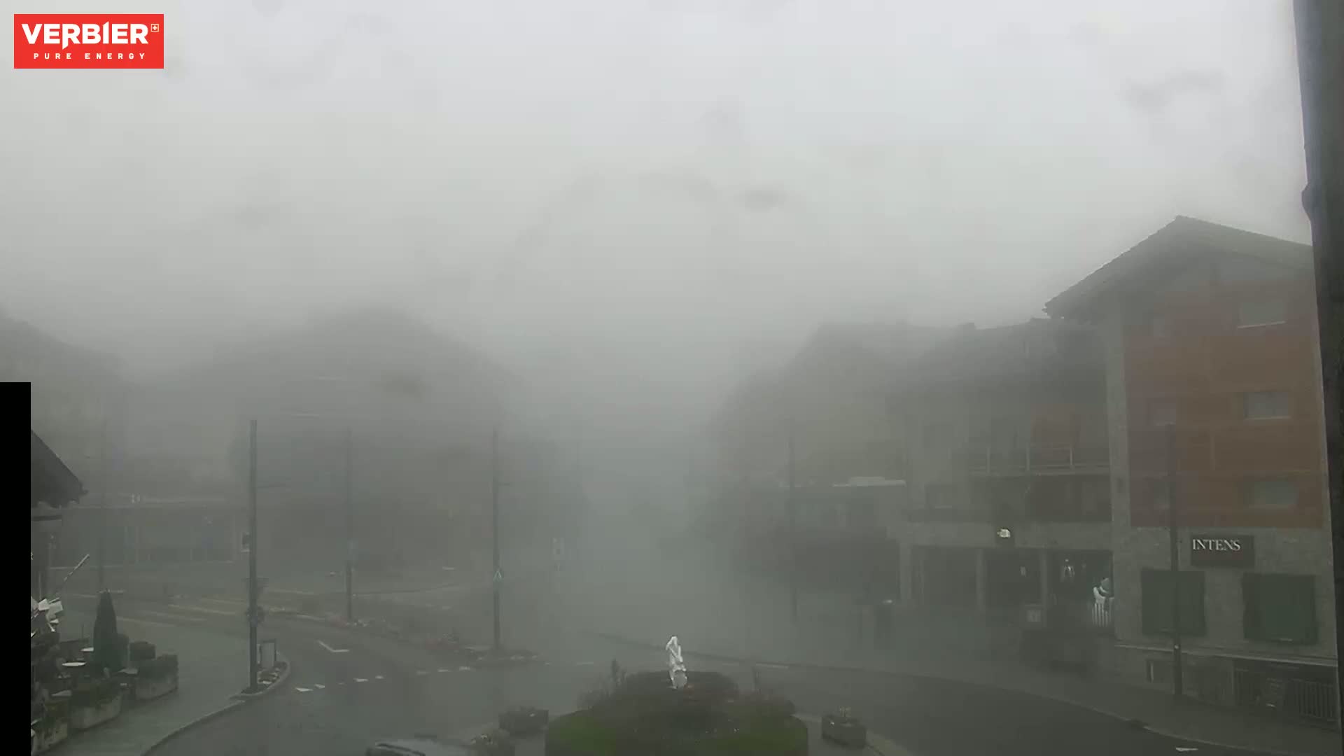 A foggy street scene shows several buildings and a small roundabout, with a statue in the center.