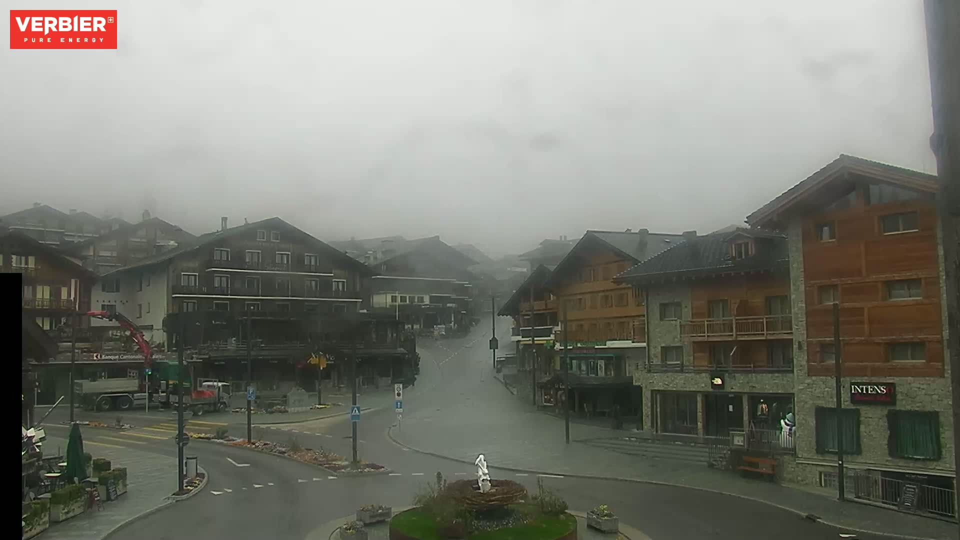 A foggy, quiet town square with several multi-story buildings is seen, with a small fountain in the center.