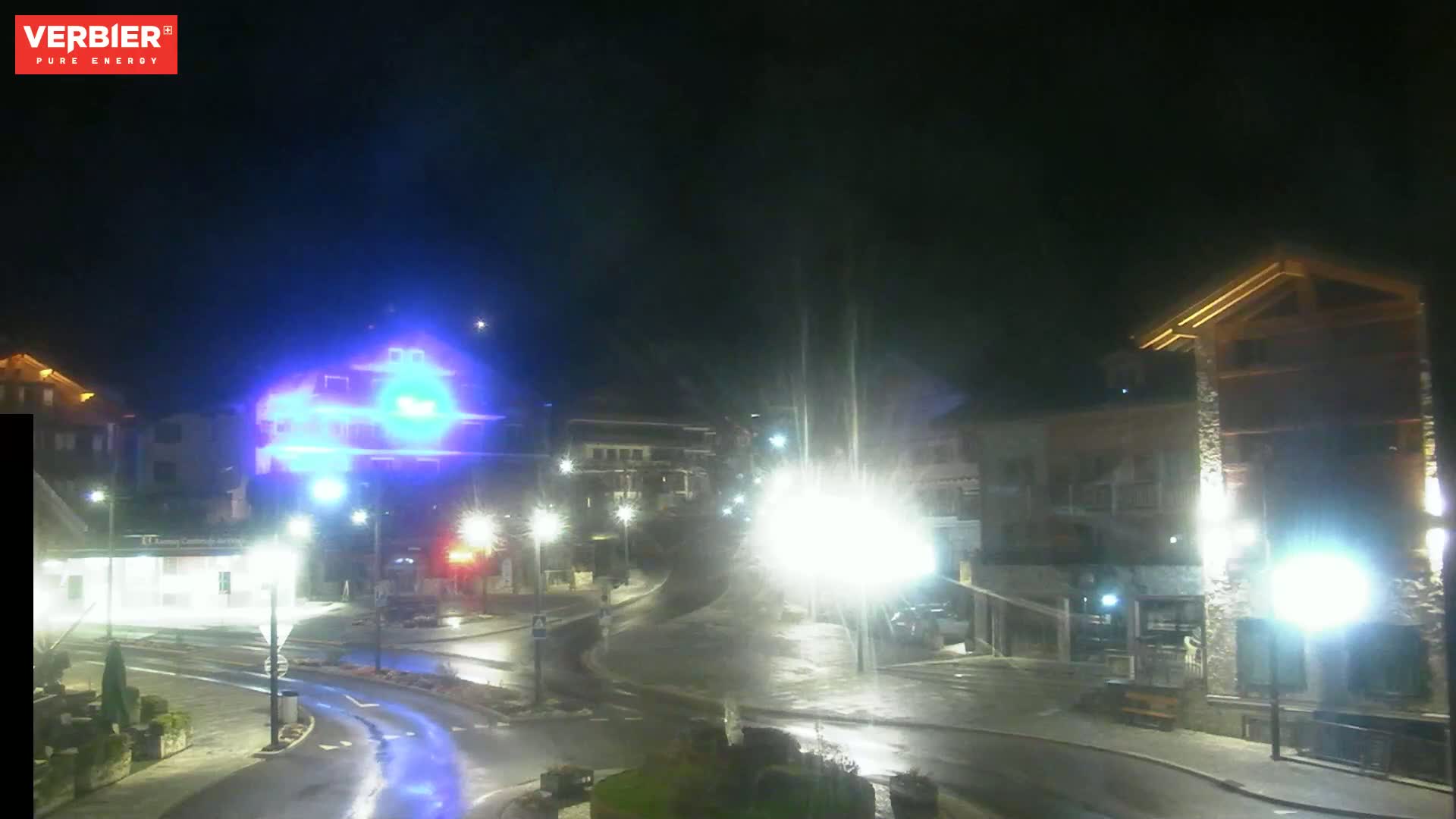 A wet, nighttime street scene shows a village square with buildings lit by streetlights.