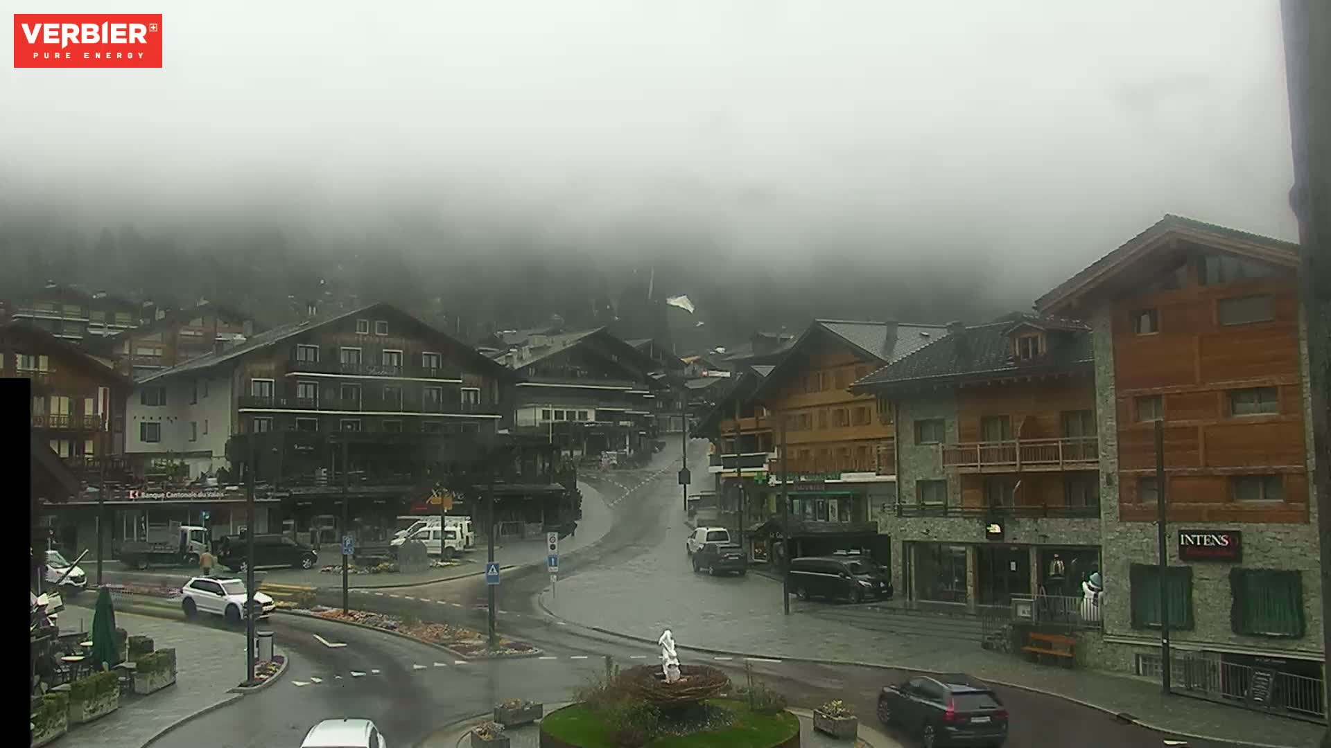 A foggy, rainy day in a village with multiple-story buildings and cars parked along the street.