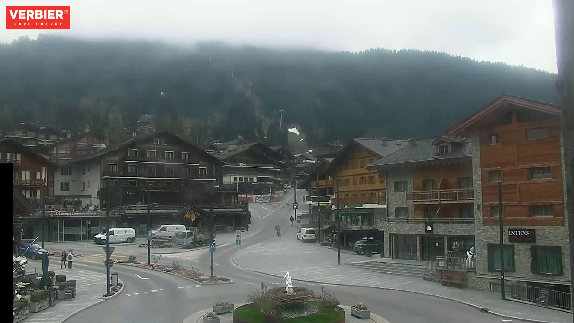 A misty mountain village scene shows several multi-story buildings surrounding a street intersection, with a few vehicles and pedestrians visible.