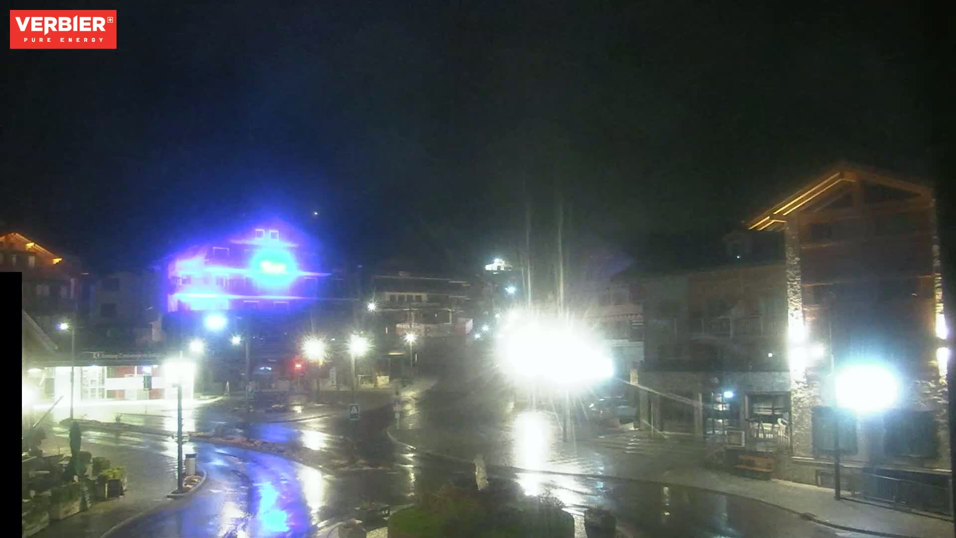 A wet, nighttime street scene shows several buildings illuminated by streetlights and other lights, with some buildings brightly lit in blue.