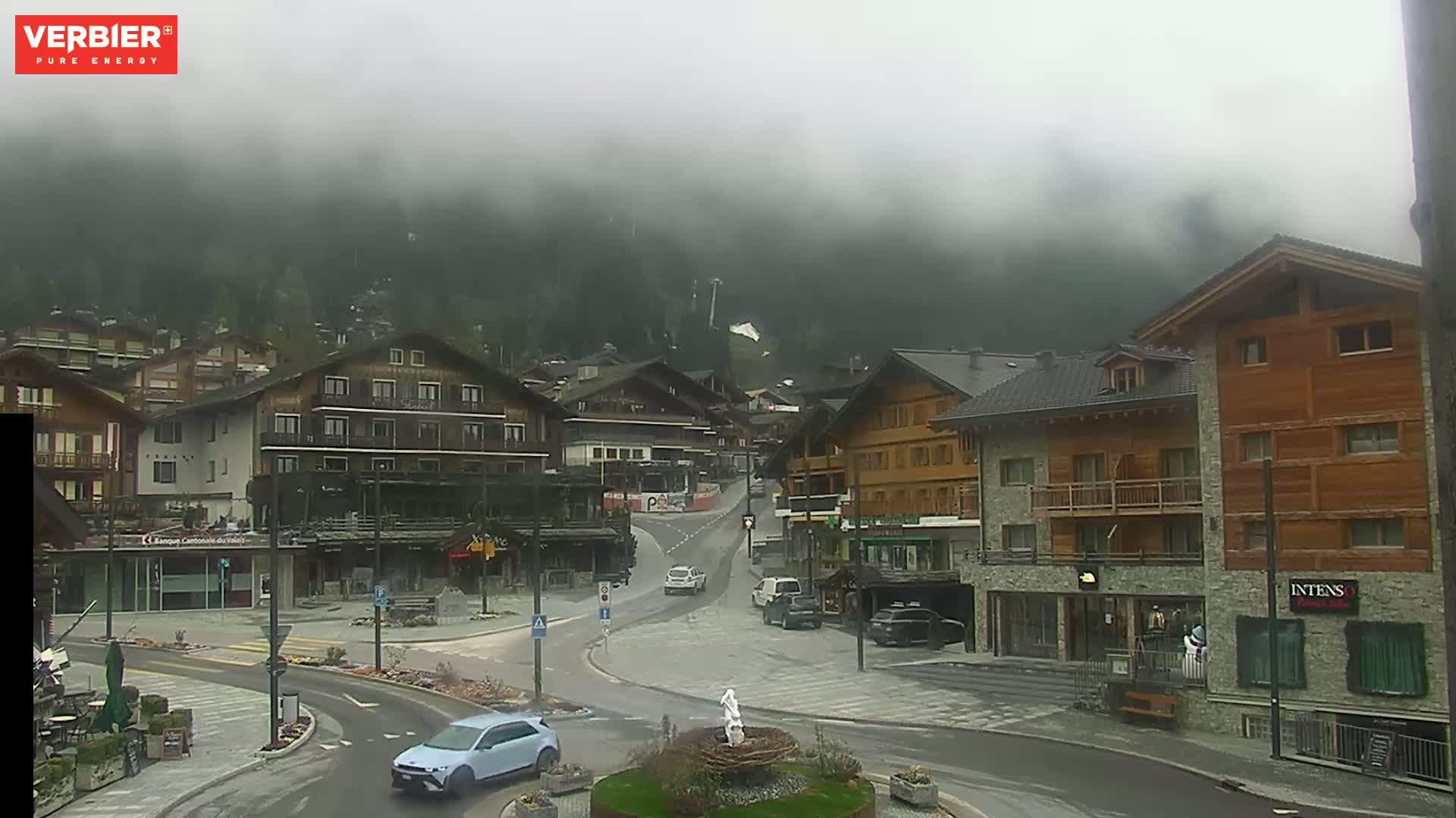 A foggy mountain village scene shows several multi-story buildings surrounding a traffic circle with a few cars and a small fountain in the center.