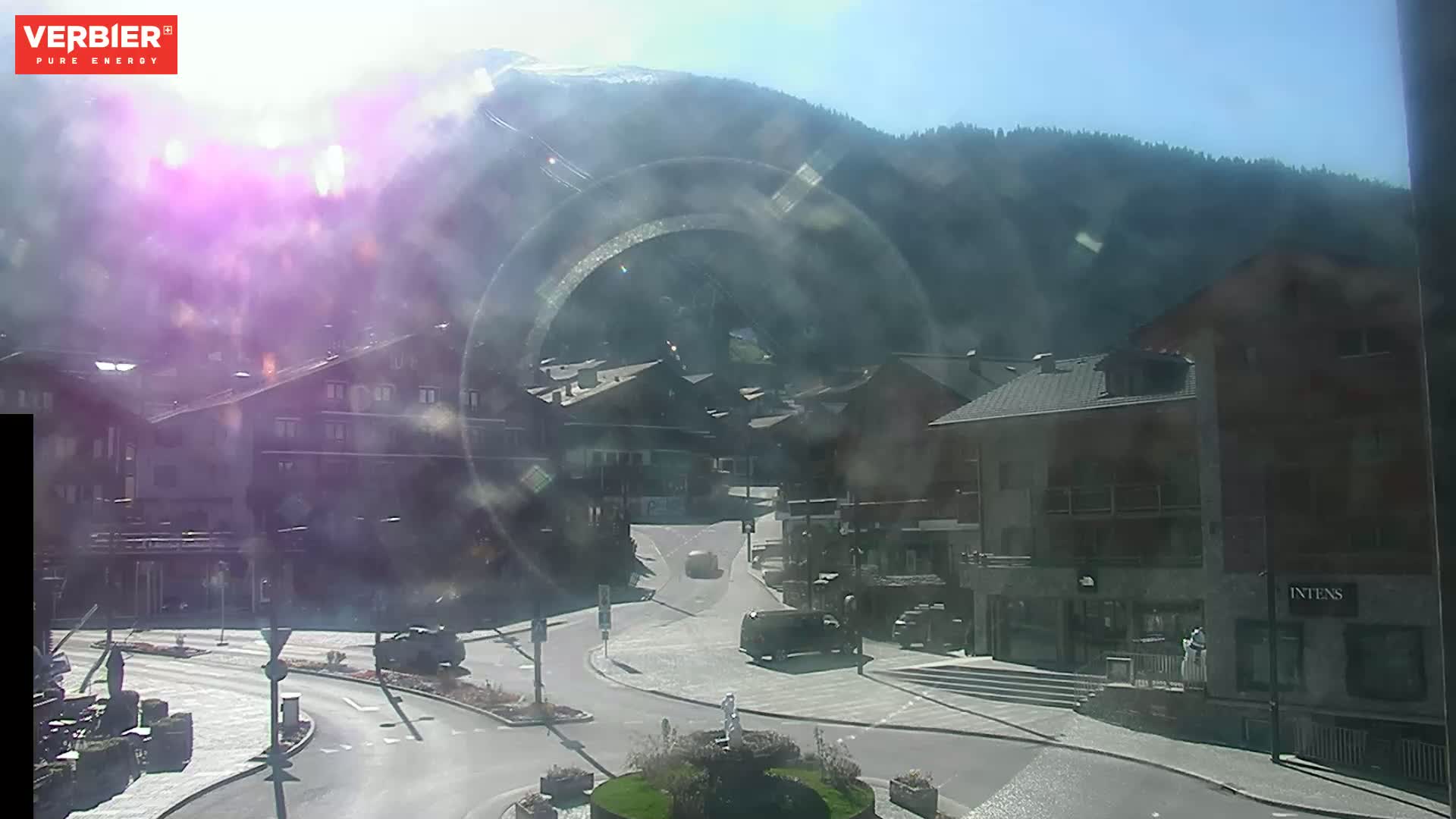 A hazy, sunny day reveals a village nestled in a valley surrounded by mountains, with cars visible on the roads.