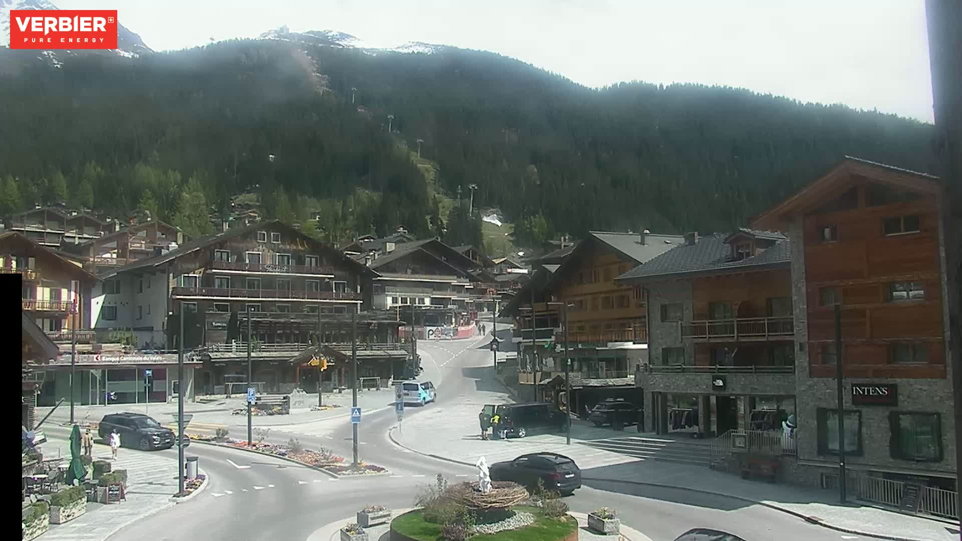 A mountain village square with several buildings, some cars, and a few pedestrians, under a mostly clear sky.