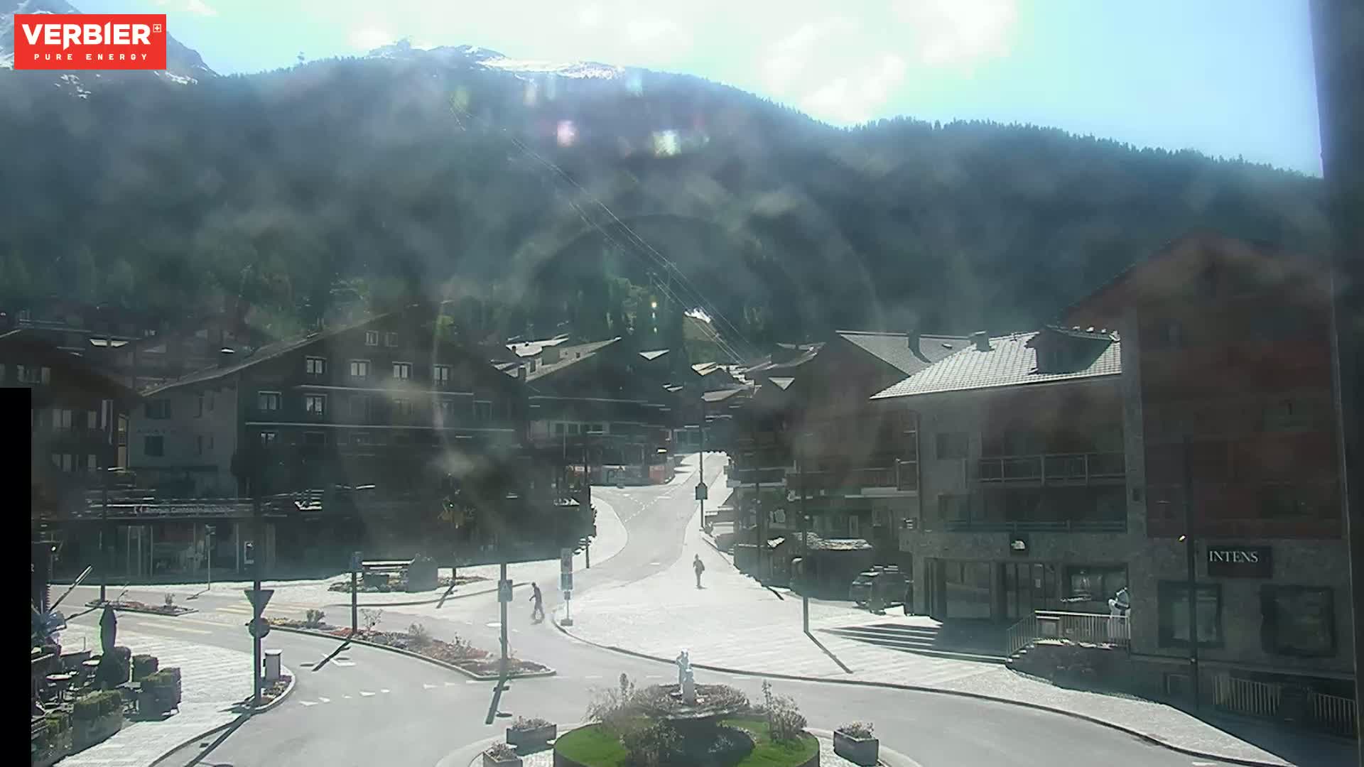 A sunny mountain village square with a few pedestrians, surrounded by buildings and a mountain in the background.