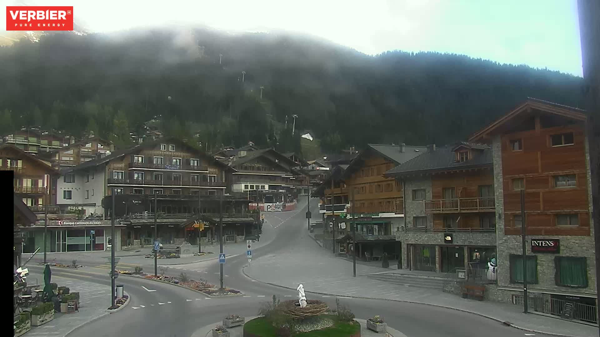 A mostly clear day reveals a mountain village with several multi-story buildings situated at the base of a mountain shrouded in mist.