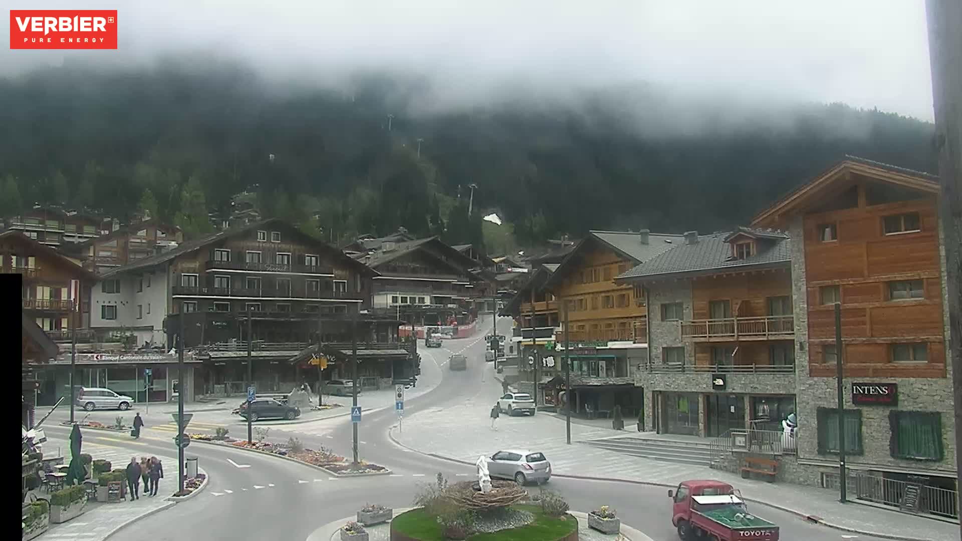 A misty mountain village square with several buildings and cars, under a cloudy sky.