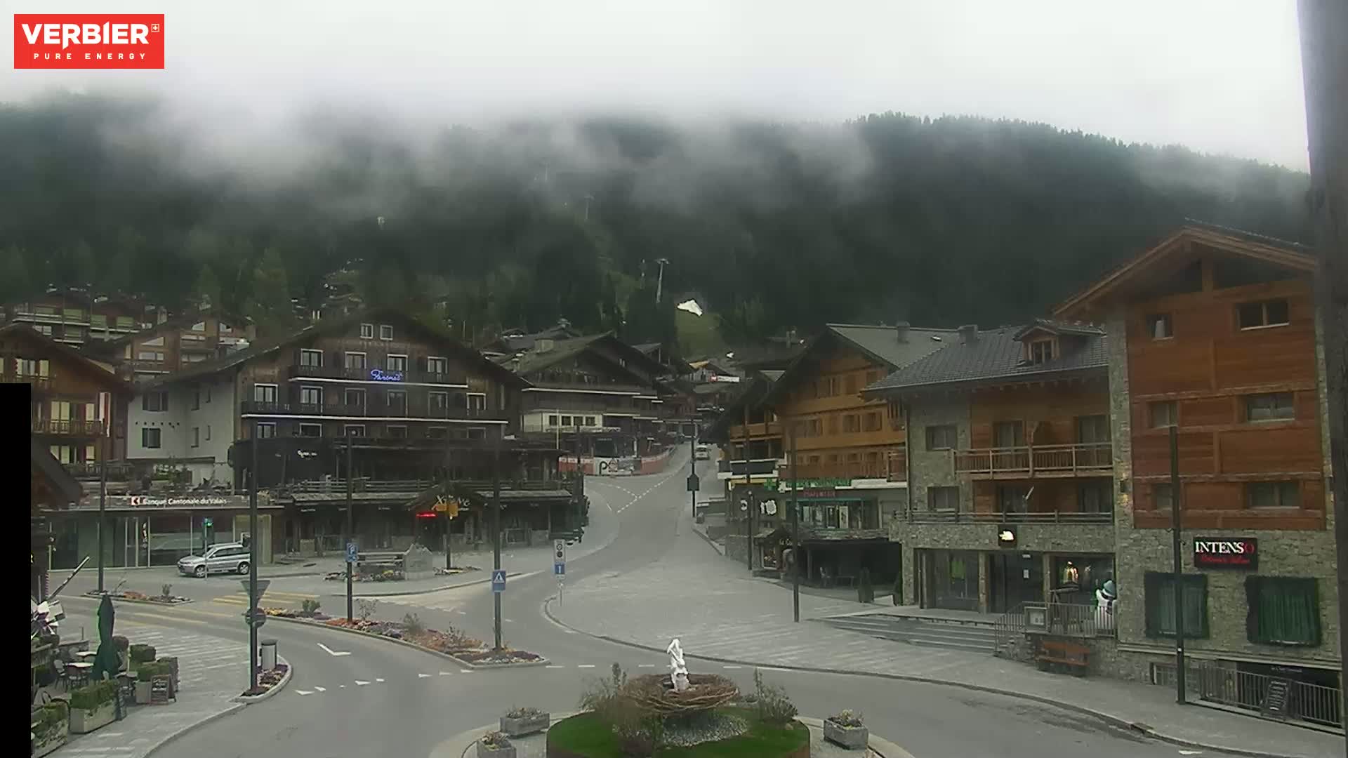 A misty mountain village square with several buildings, a small fountain, and relatively empty streets.