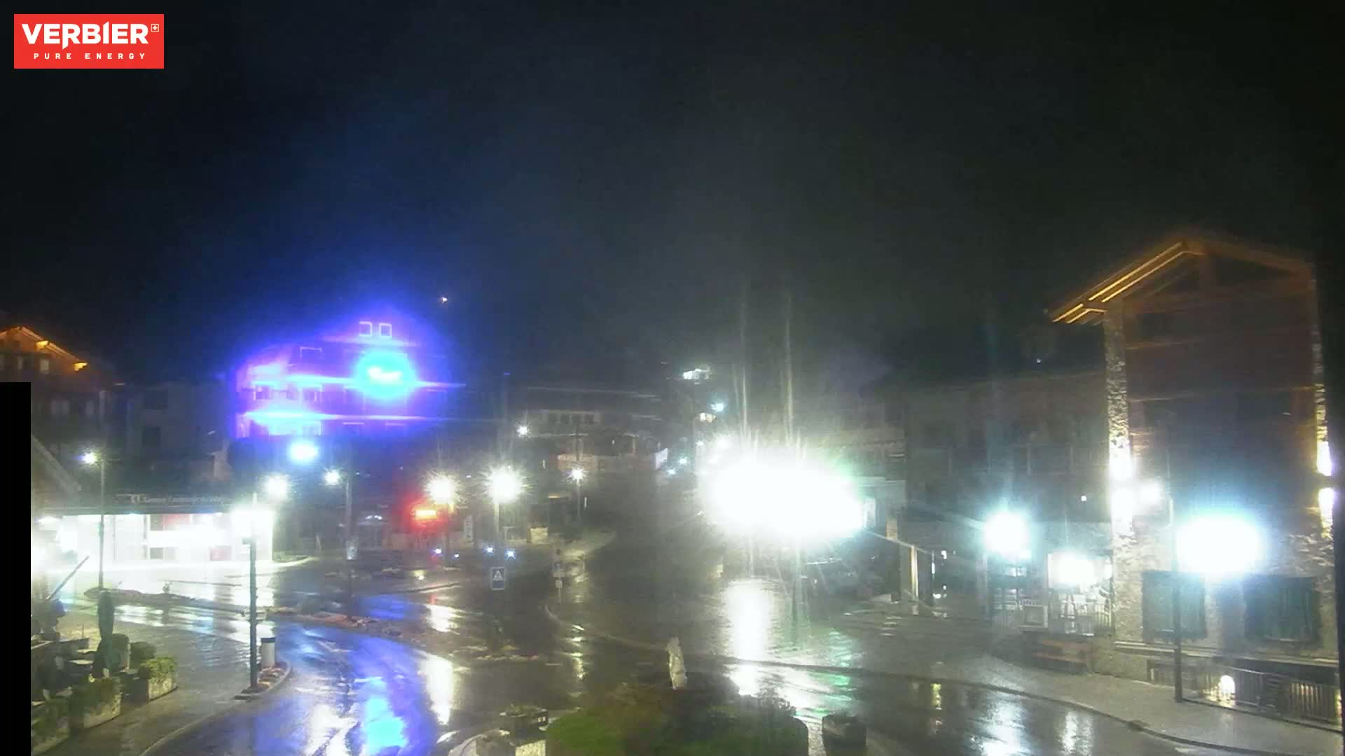 A rain-slicked town square at night, illuminated by streetlights, with several buildings visible.