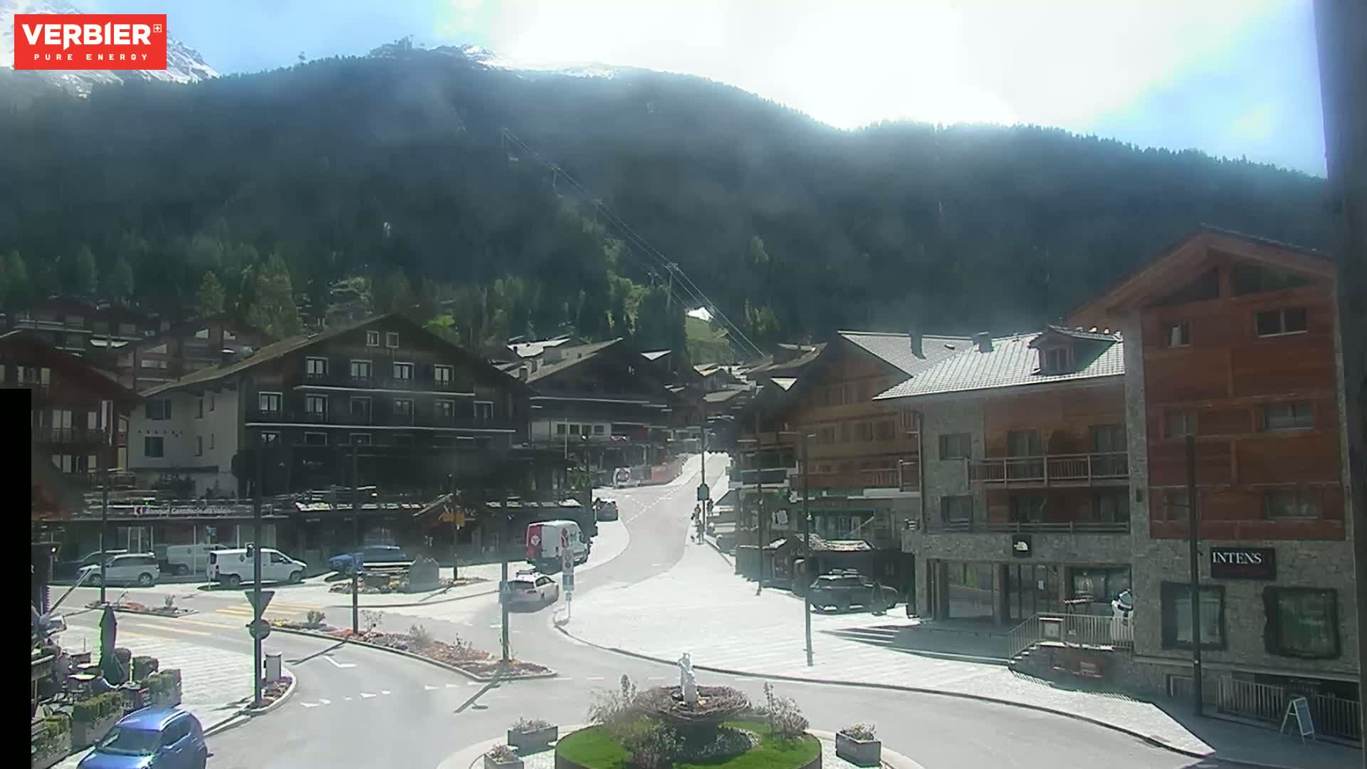 A sunny mountain village scene shows several buildings, cars, and a roundabout, with a mountain range visible in the background.