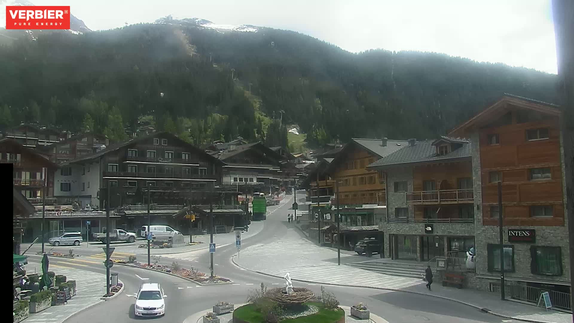 A mountain village with wood-framed buildings and a few cars is visible on an overcast day, with a mountain range in the background showing some patches of snow.
