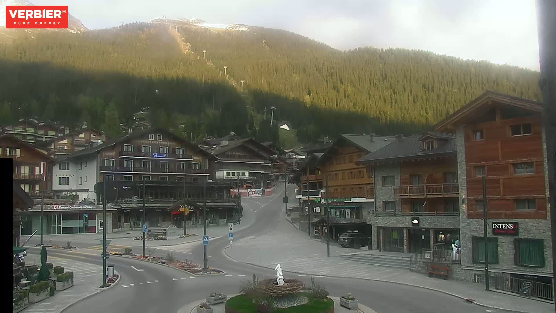 A mostly clear day reveals a mountain village nestled at the base of a forested mountain, with a few buildings lining a quiet street.