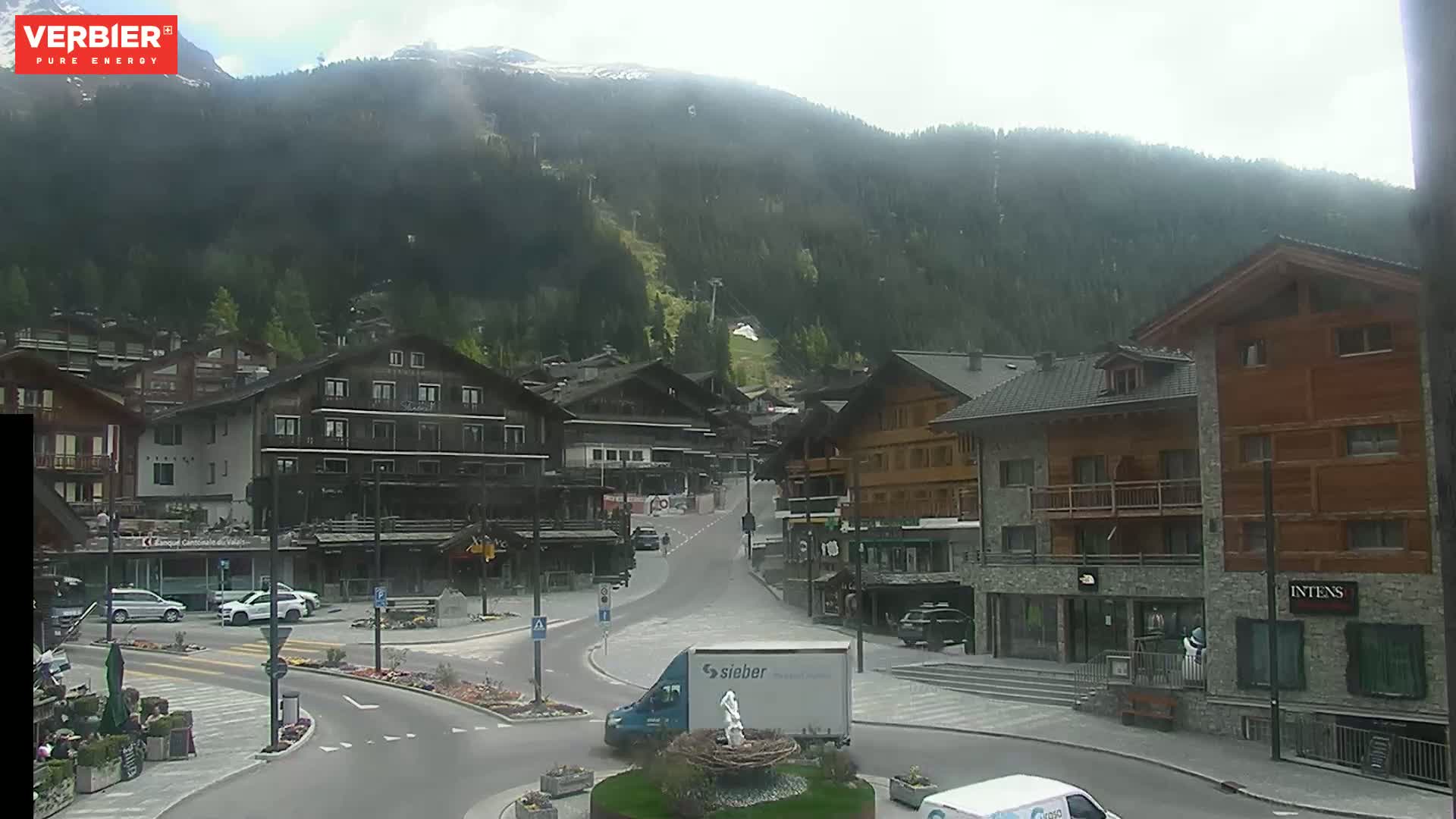 A mountain village square with several buildings, a few cars, and a delivery truck is visible under an overcast sky.