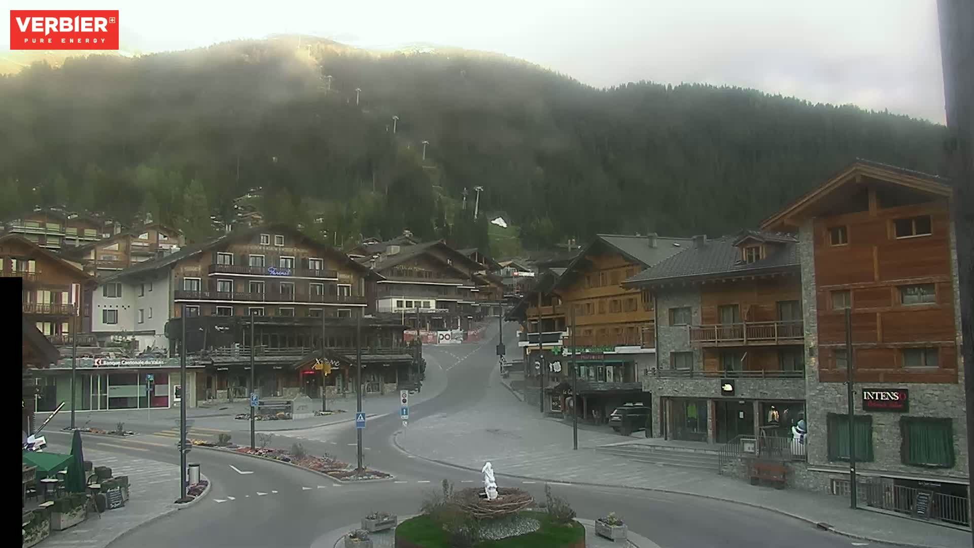 A mostly clear view of a village nestled in a valley, with a mountain shrouded in fog in the background.
