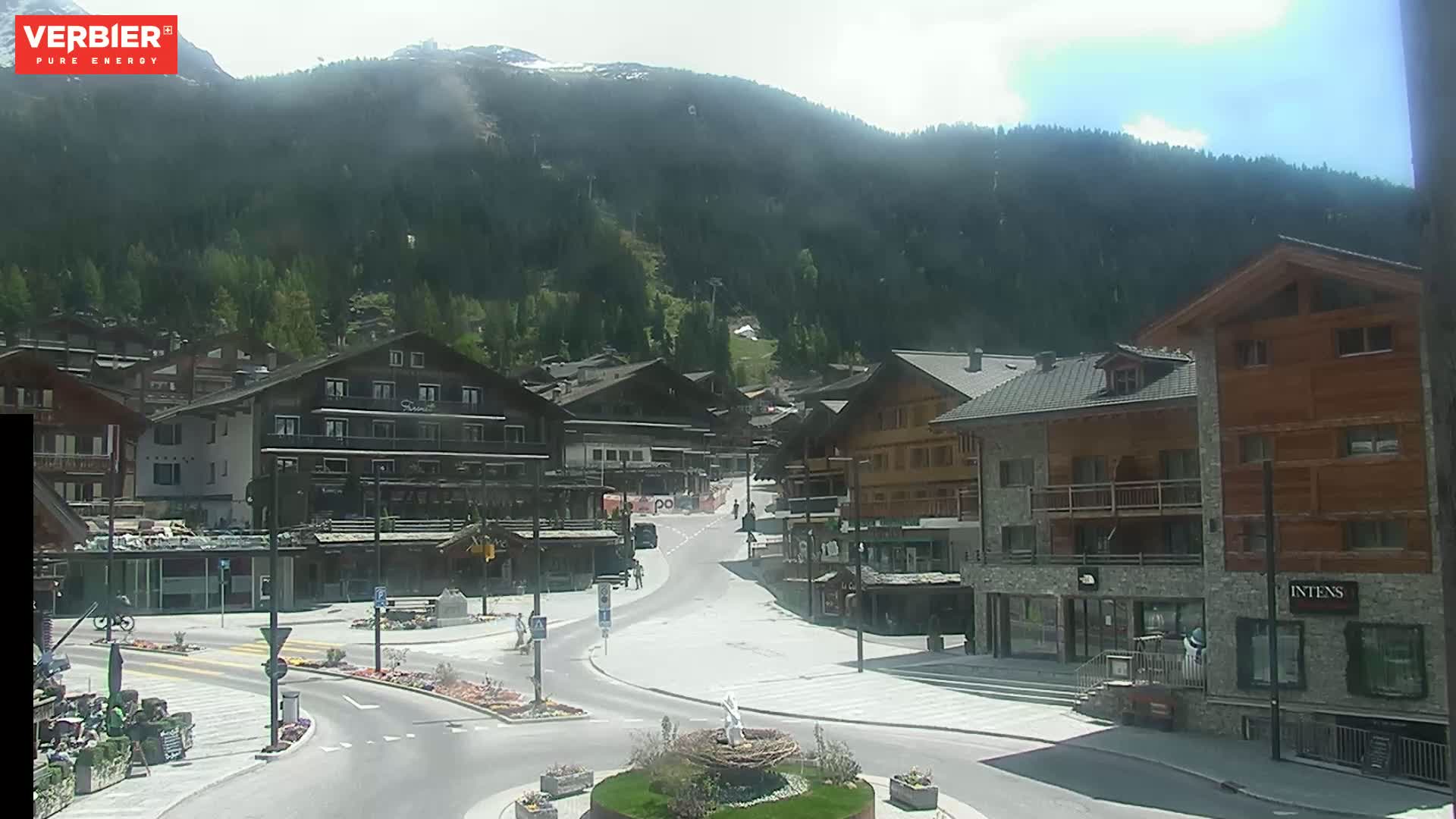 A mostly sunny view of a village nestled in a valley surrounded by mountains, with a mostly empty town square and roads.