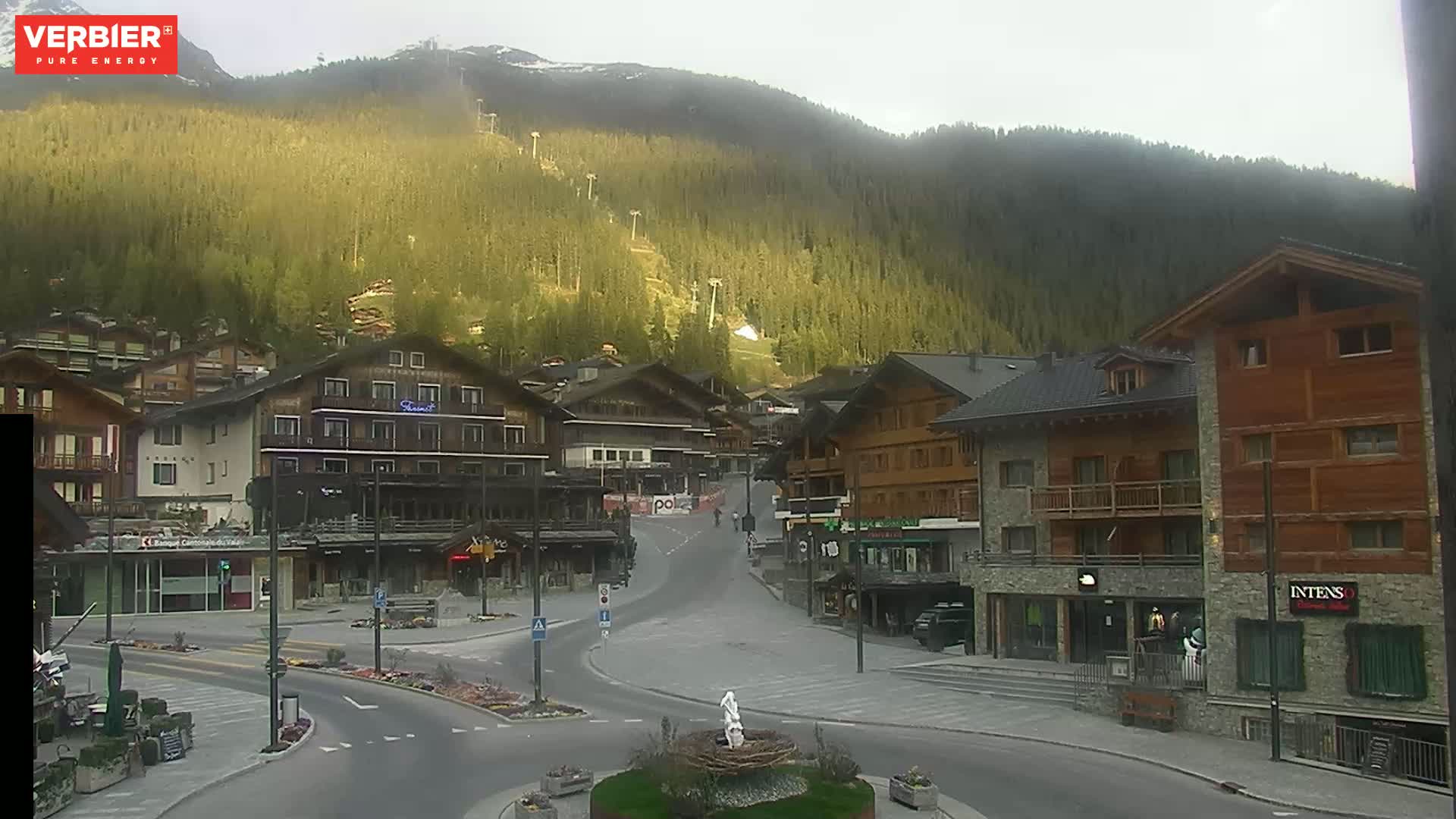 A mostly clear, hazy day reveals a mountain village nestled at the base of a steep, forested mountain with ski lifts visible.
