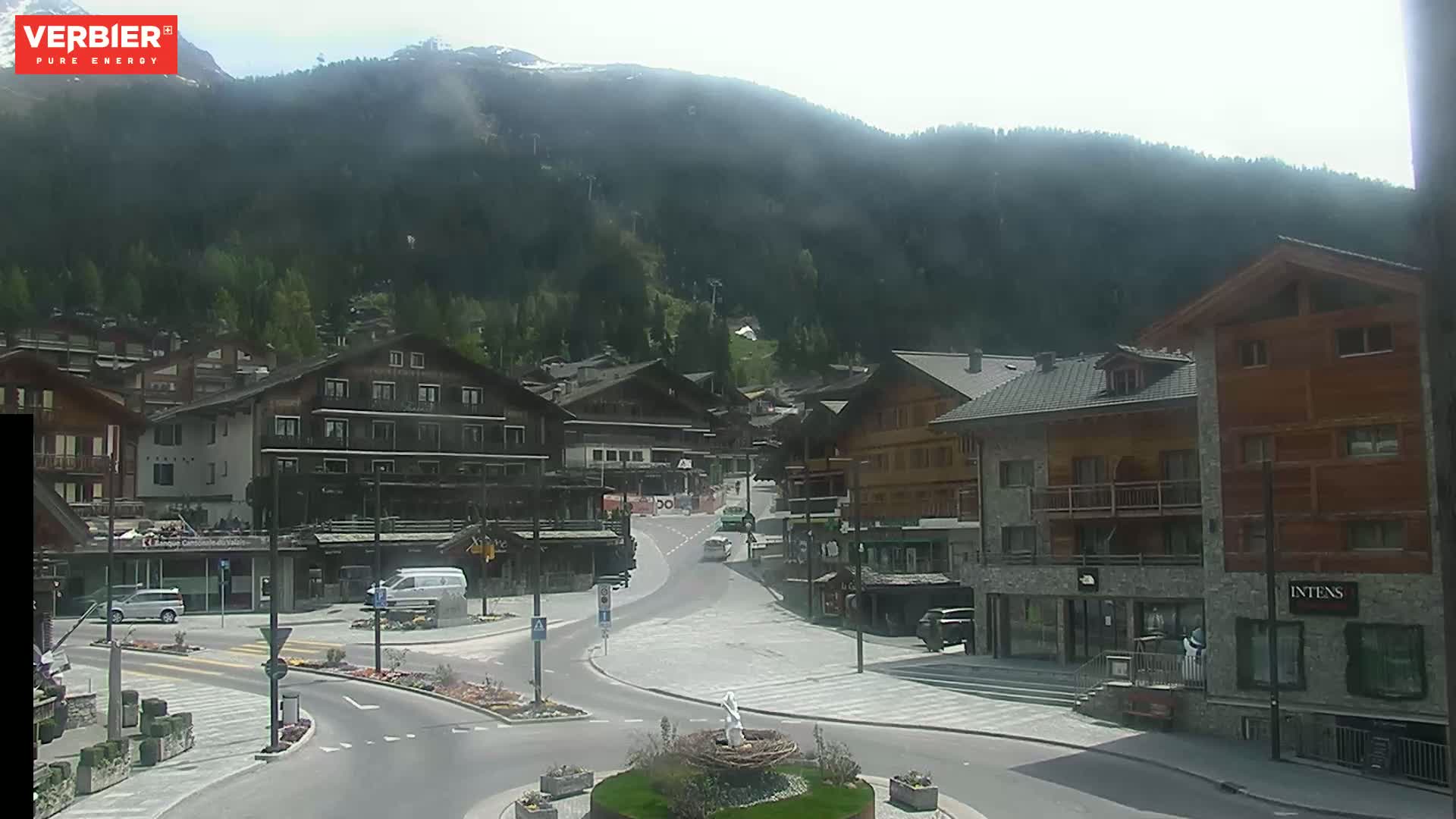 A hazy view of a town nestled in a valley surrounded by mountains on a partly sunny day.