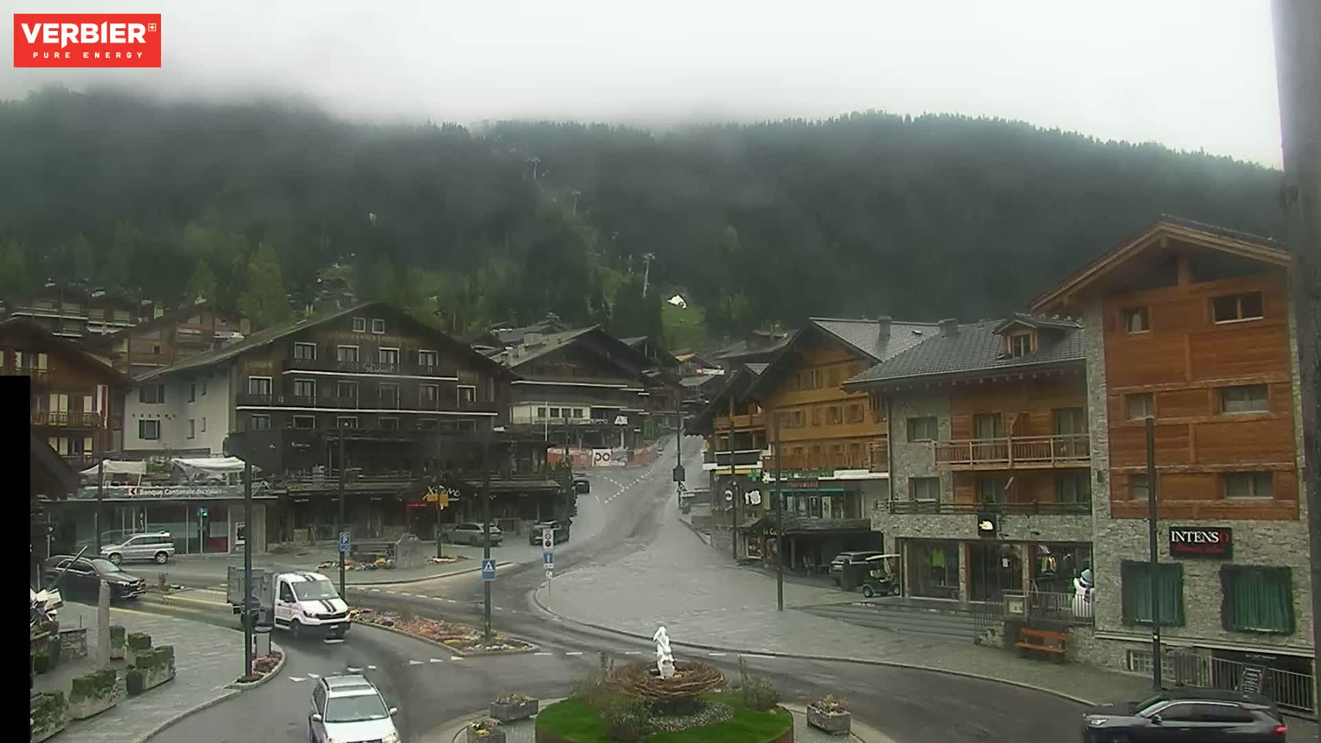 A misty mountain village with several multi-story buildings, cars parked and driving on wet roads, and a small fountain in a roundabout.