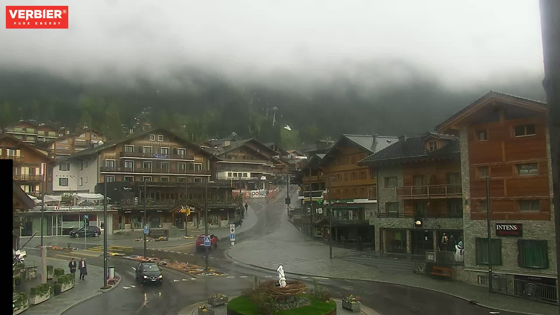 A wet, foggy mountain town square with several multi-story buildings, cars, and a few pedestrians.