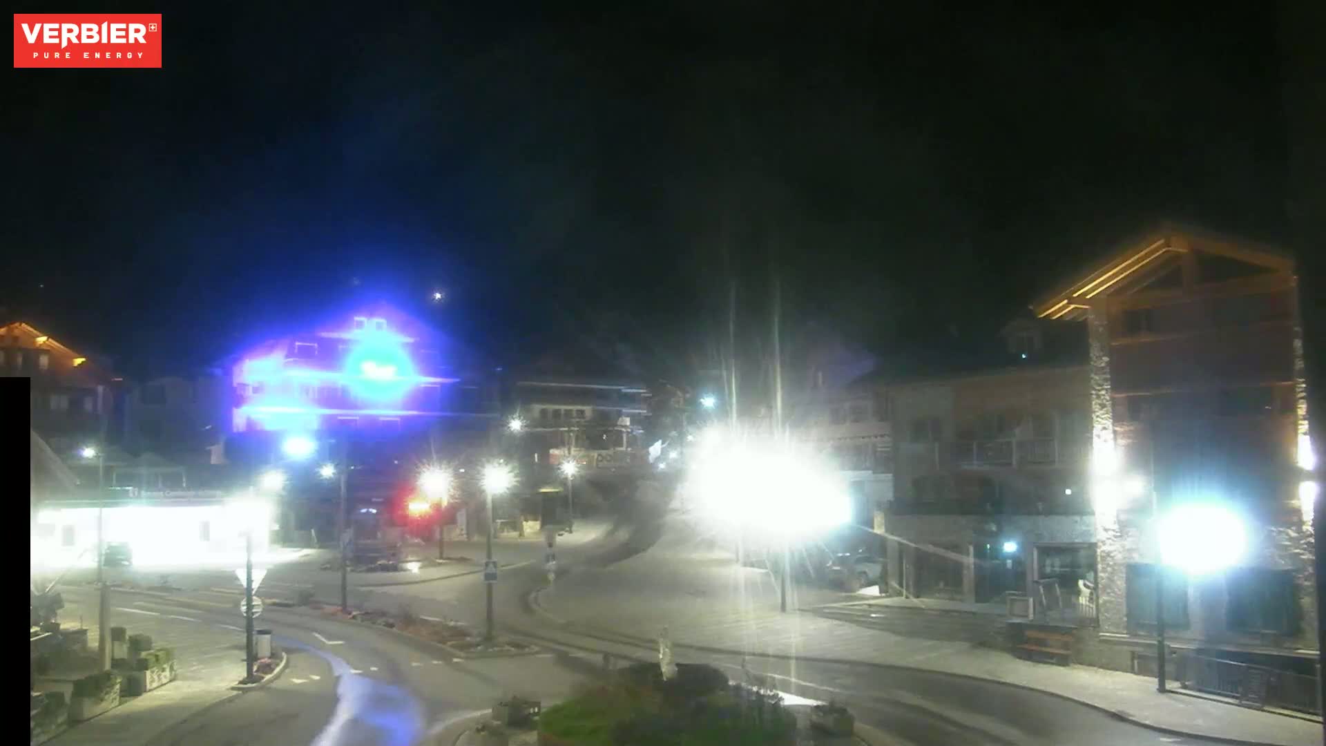 A nighttime, brightly lit town square with several buildings, streetlights, and a few vehicles.