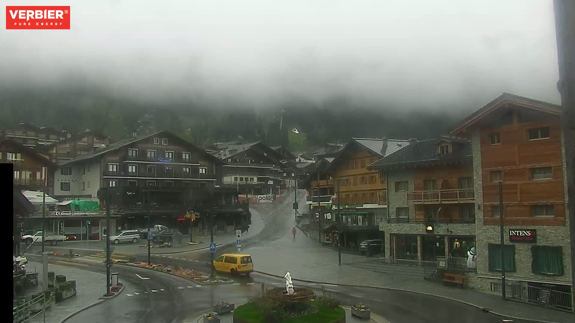 A foggy, wet town square in a mountain village, with several multi-story buildings surrounding a wet road.