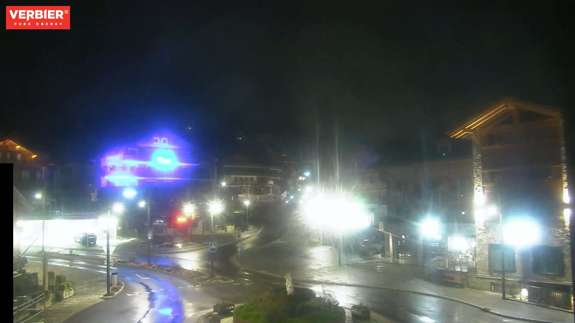 A wet, nighttime street scene shows a town square with several buildings, streetlights, and vehicles.