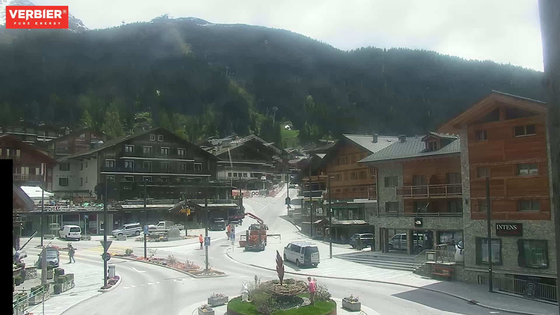 A mostly sunny day in a mountain village with several buildings, vehicles, and people visible in a town square.