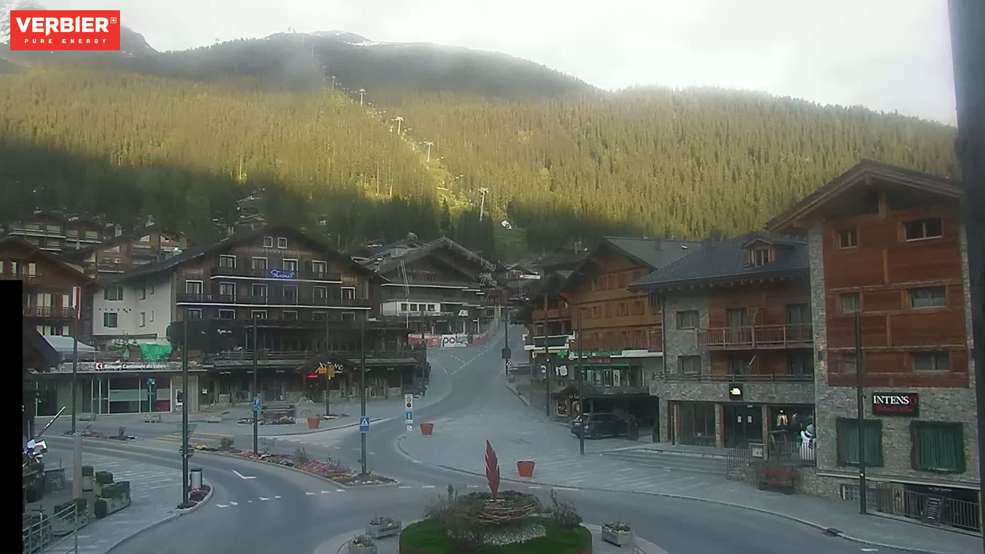 A mostly empty town square in a mountain village, with several multi-story buildings lining the streets, under an overcast sky.