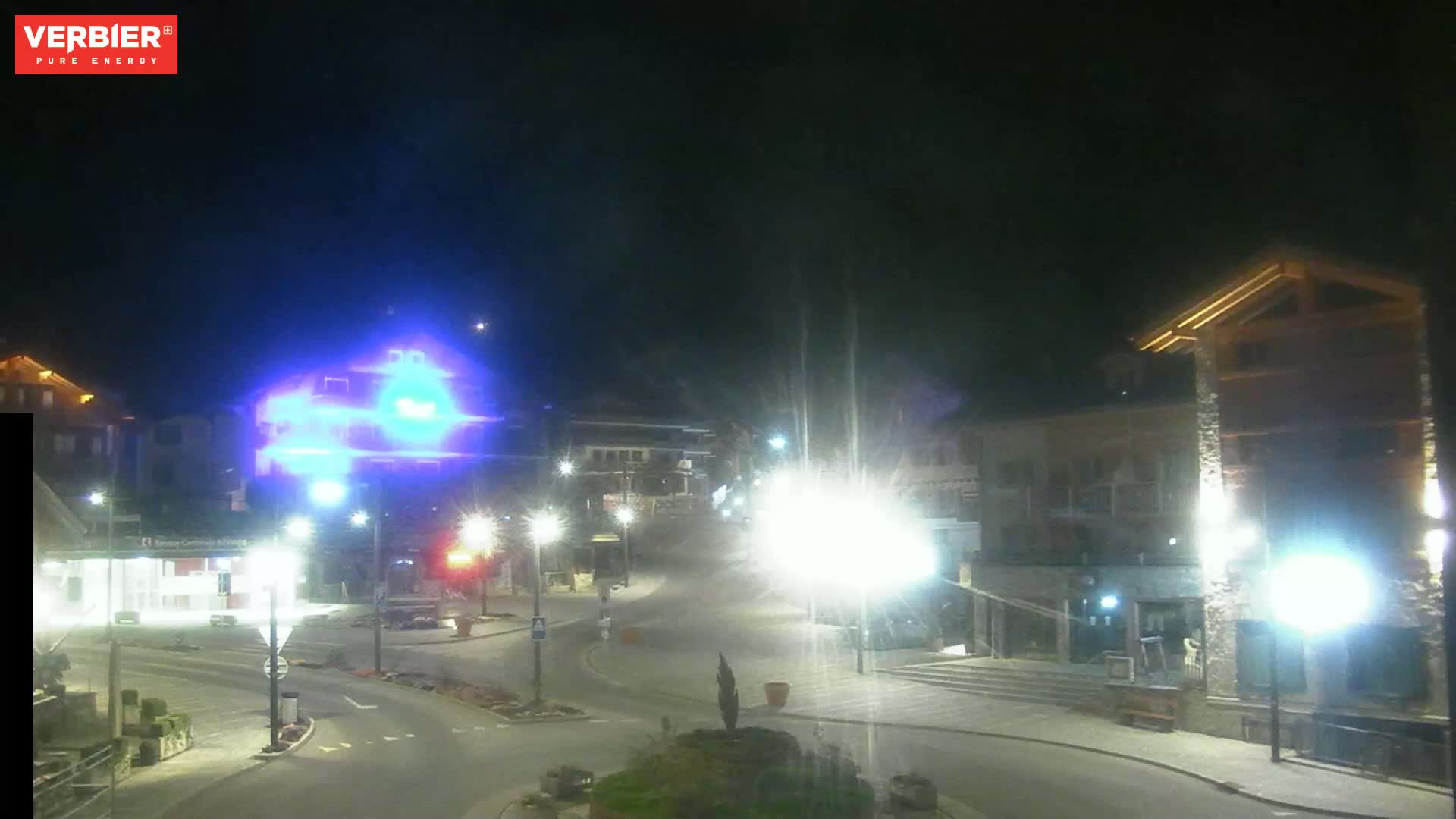 A nighttime view of a town square with buildings illuminated by streetlights under a clear, dark sky.