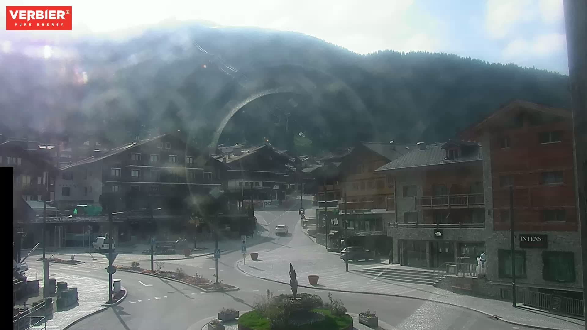 A hazy, sunny day reveals a village nestled in a valley, surrounded by mountains.