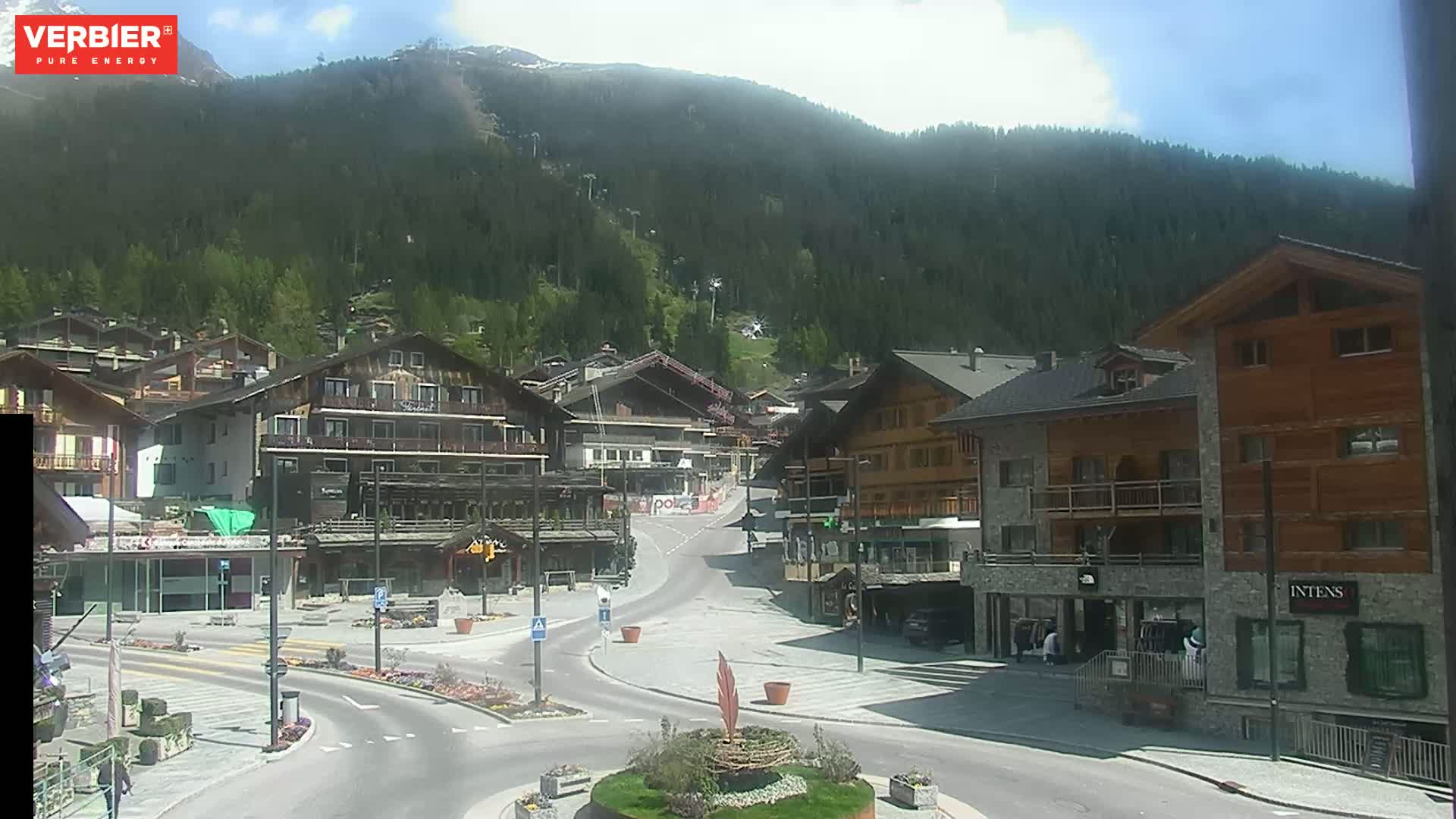 A sunny mountain town square with several buildings, a paved road, and a grassy roundabout is nestled against a backdrop of green, tree-covered slopes.