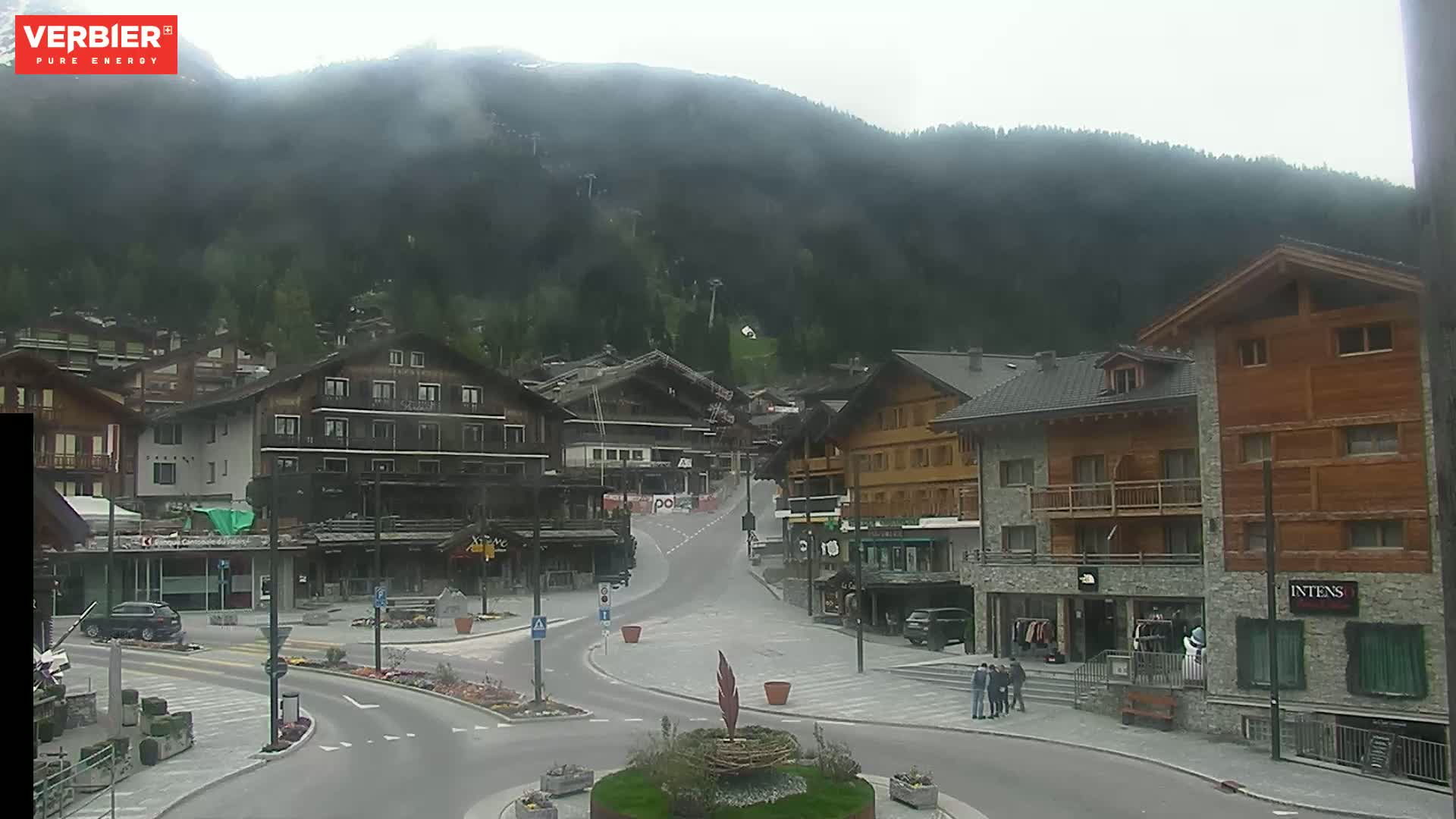 A mostly clear view of a town square nestled in a valley between forested, misty mountains.