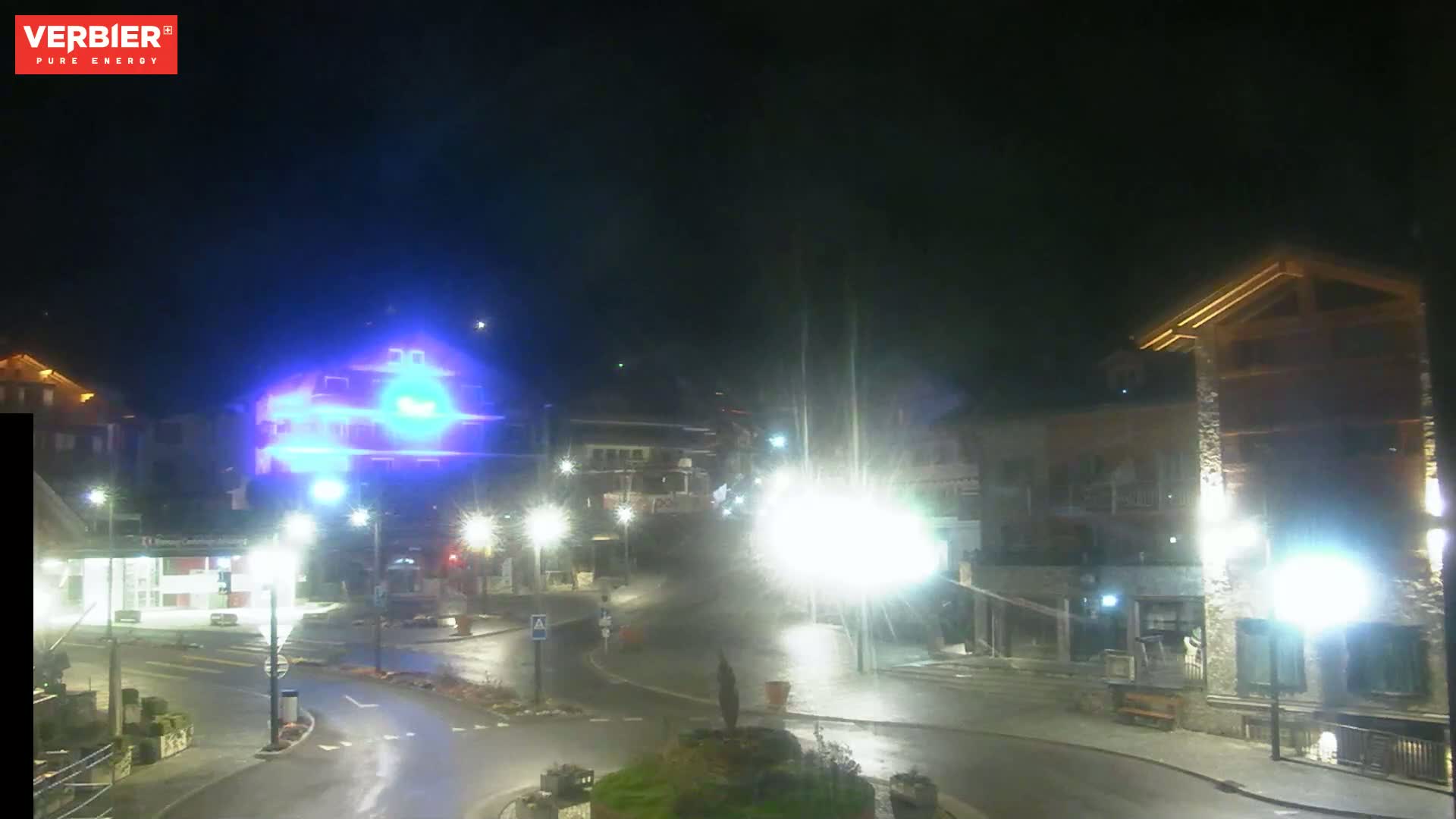 A wet, nighttime street scene shows a roundabout in a village with brightly lit buildings and streetlights.