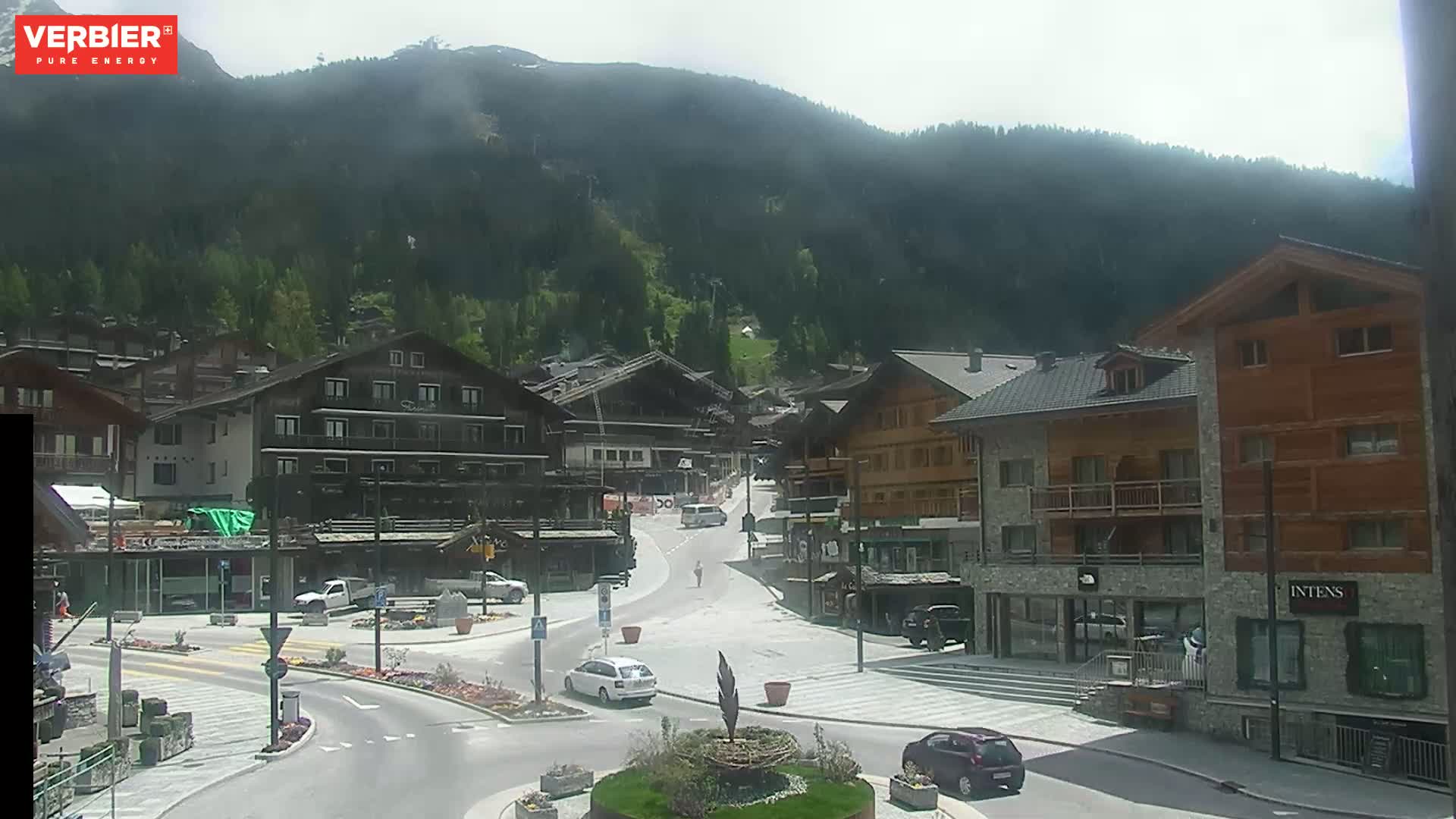 A mountain village square with several buildings, a few cars, and a person walking, under a mostly cloudy sky.