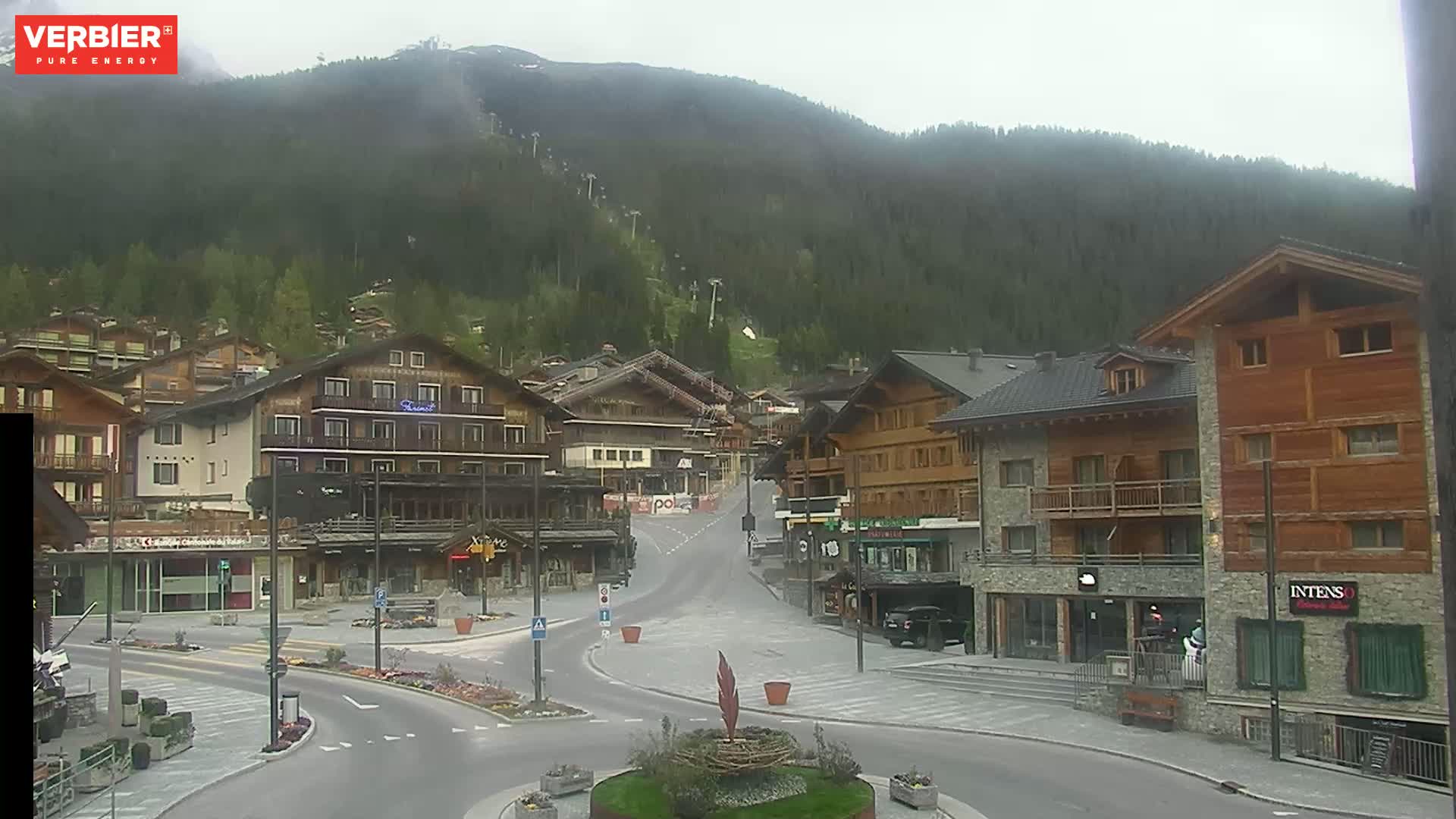 A mostly empty town square in a mountain village on an overcast day, with several chalet-style buildings lining the street, leading towards a forested hillside with ski lifts visible in the distance.
