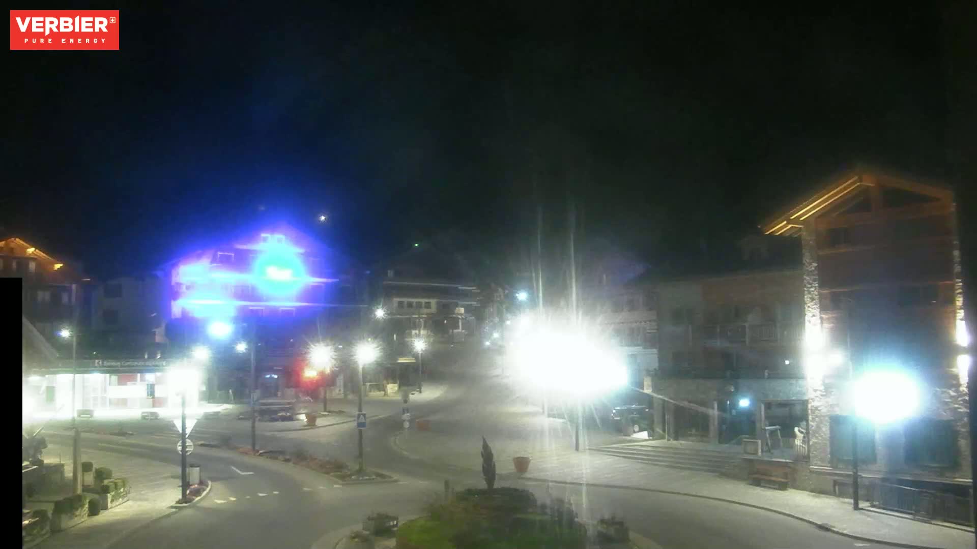 A nighttime view of a town square with brightly lit buildings and streets, under a clear, dark sky.