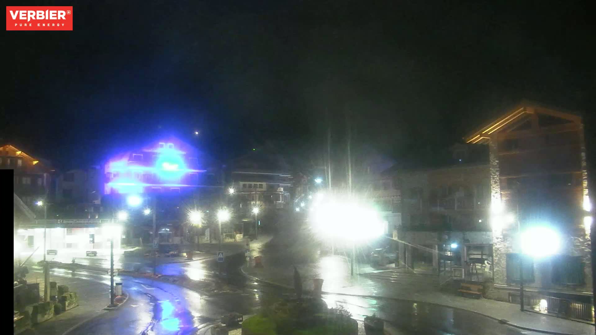 A wet, nighttime scene shows a village square with several buildings illuminated by streetlights, and a building in the background is lit in bright blue.