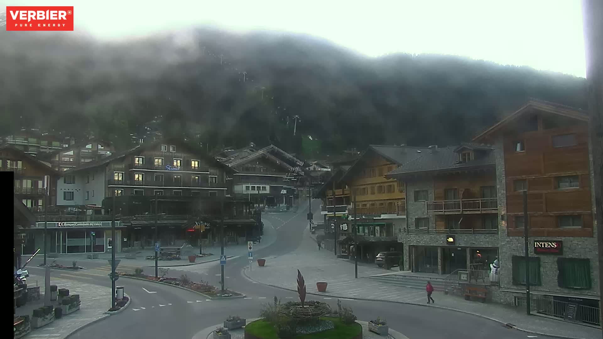 A misty mountain village square with several buildings, a roundabout, and a few pedestrians.