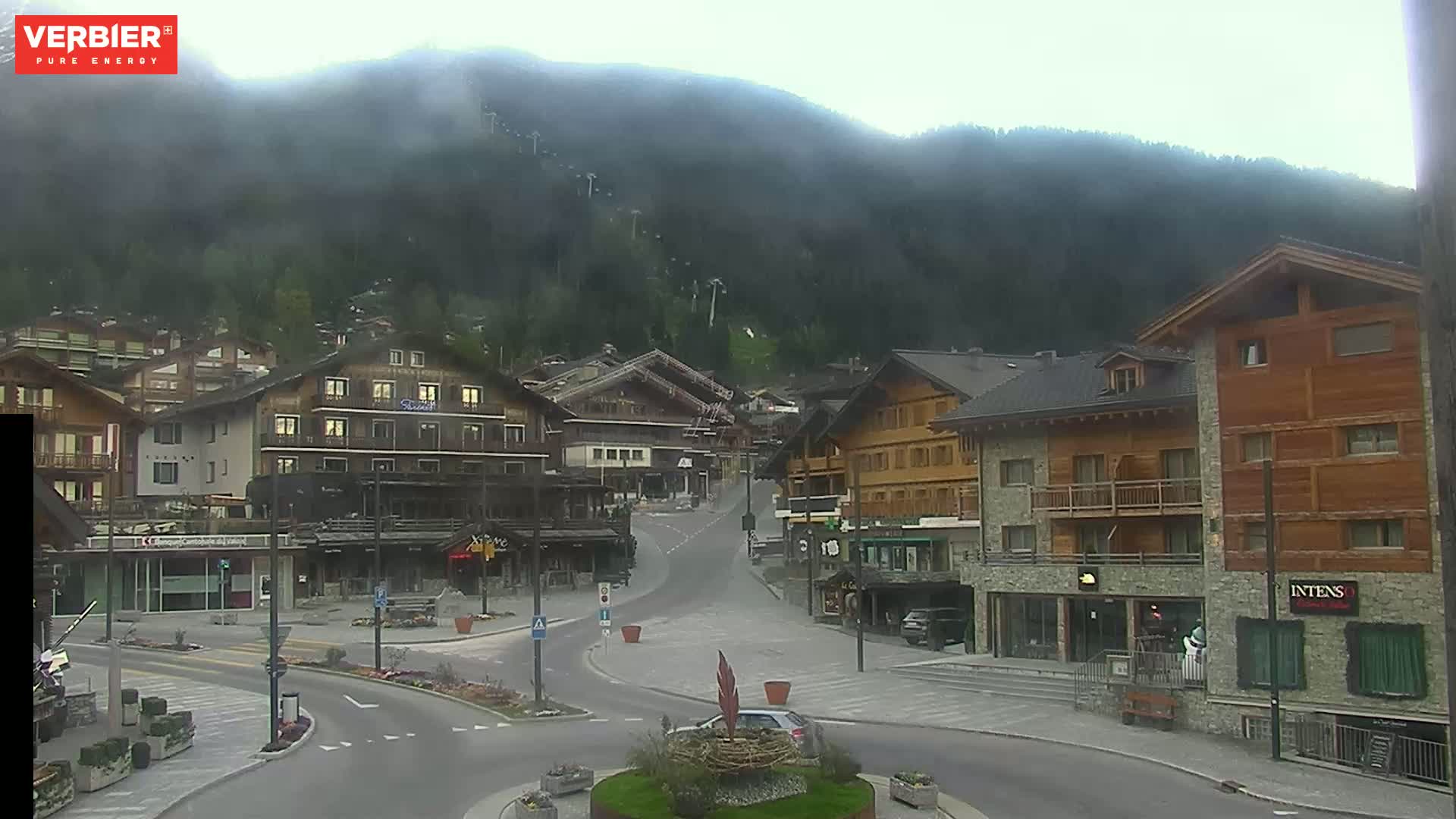 A mostly clear, misty day reveals a mountain village with chalet-style buildings and quiet streets, nestled at the base of a large, forested mountain.