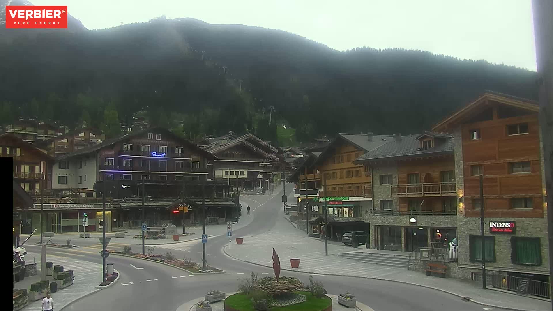 A mostly empty town square in a mountainous area on an overcast day, with several multi-story buildings lining the streets.