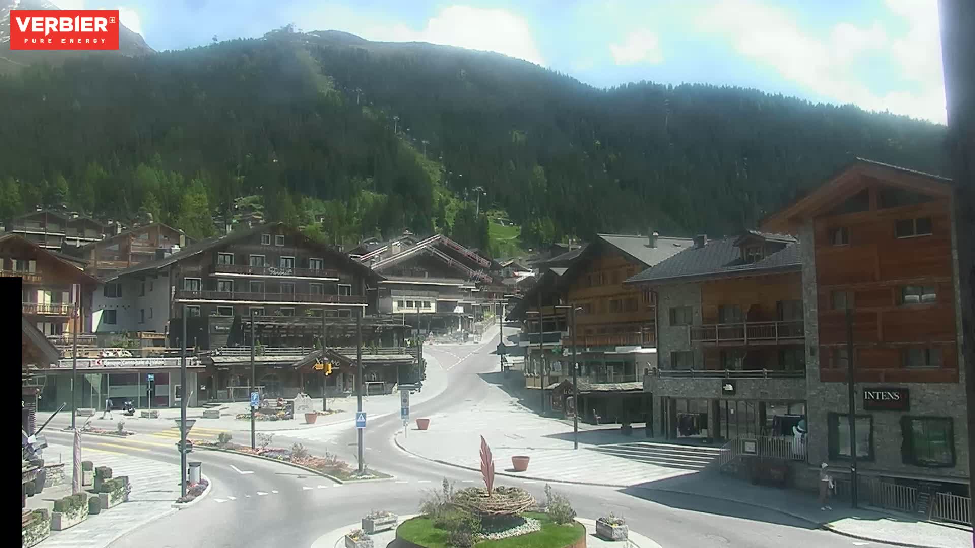 A sunny mountain village scene shows a plaza with several buildings and roads, nestled at the base of a verdant mountain.