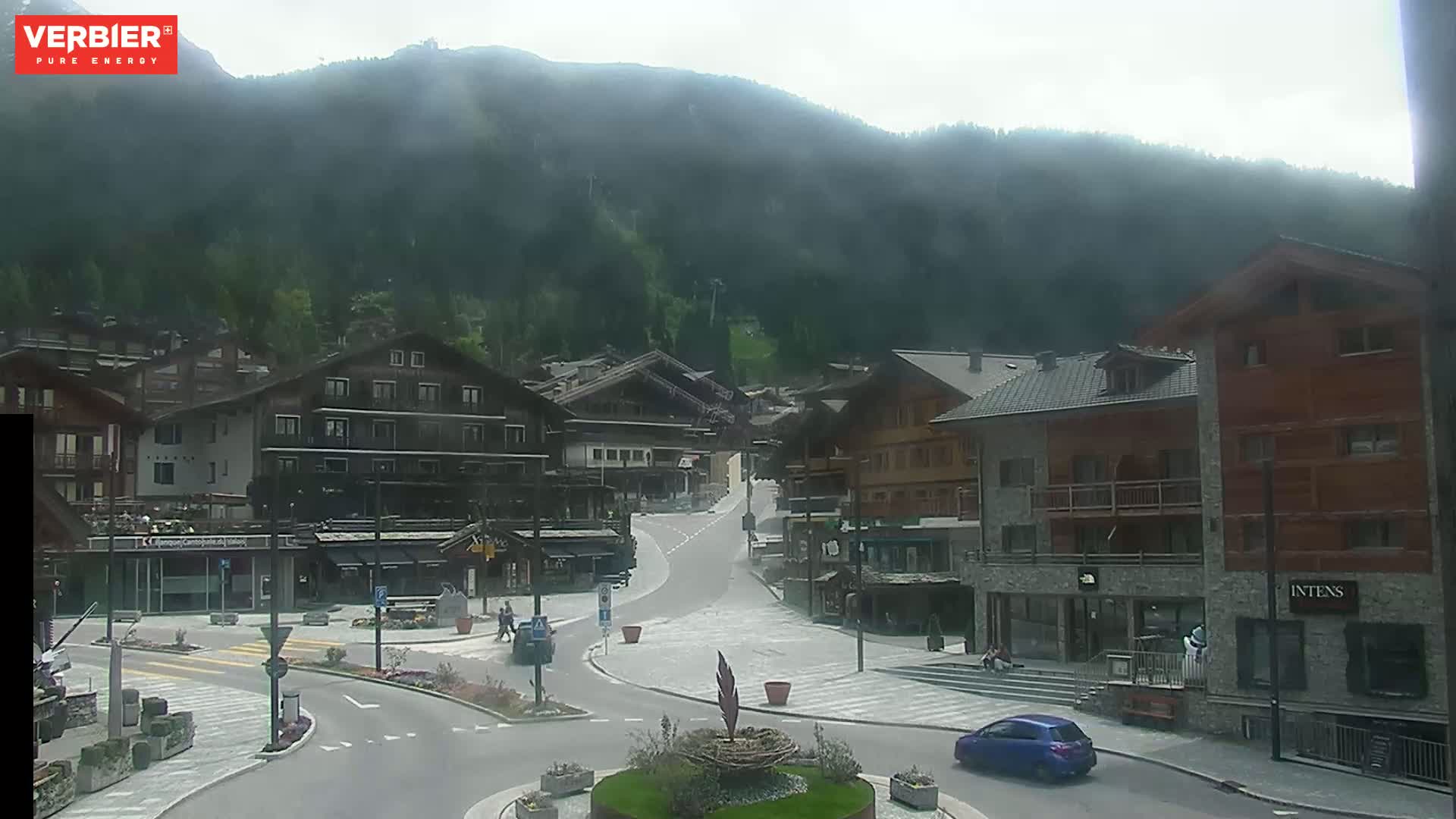 A mostly clear day shows a village nestled in a valley, surrounded by a mountainside covered in green trees and a few buildings.