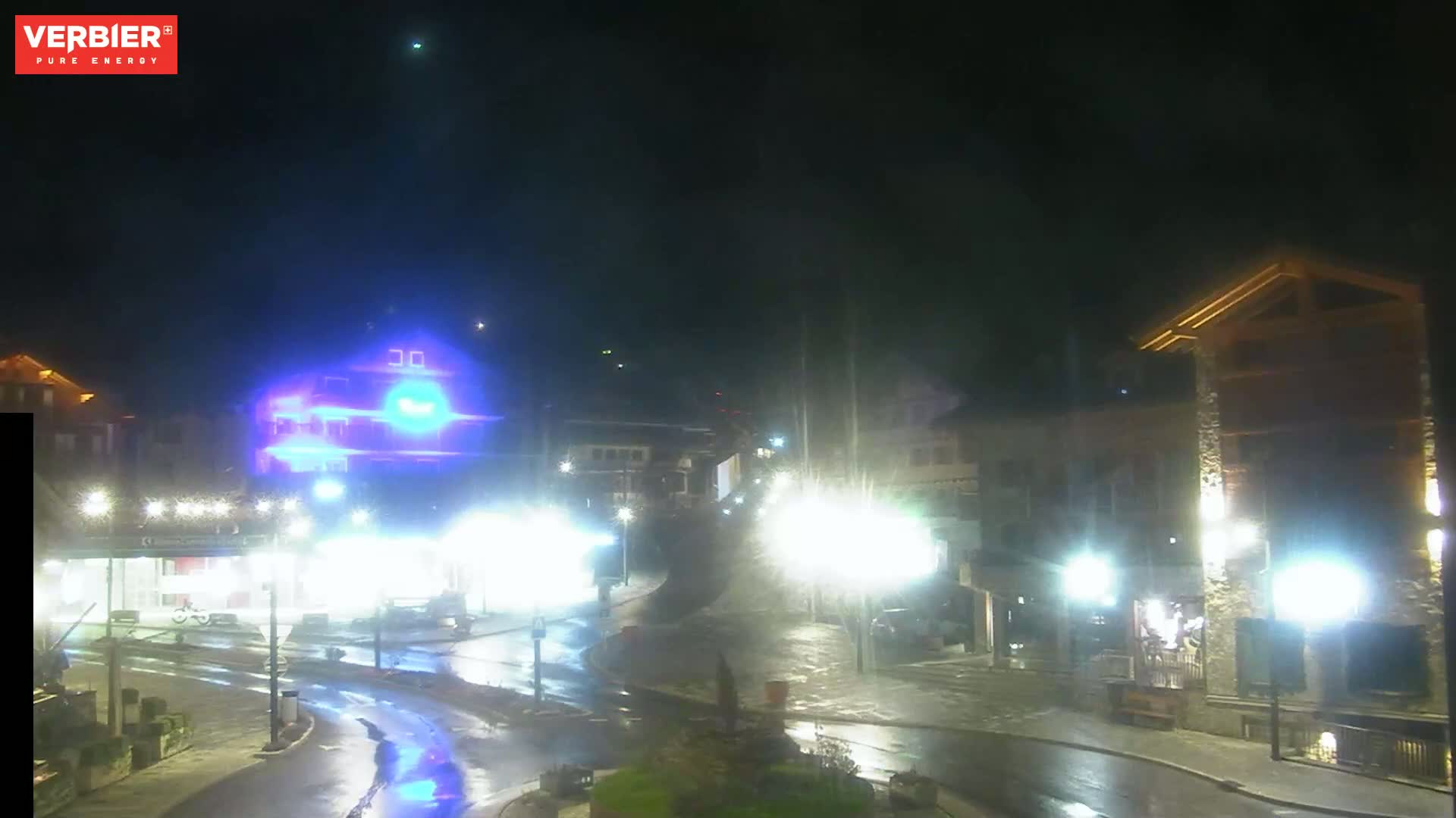 A wet, nighttime street scene shows brightly lit buildings and streetlights reflecting on a dark, rain-slicked road.
