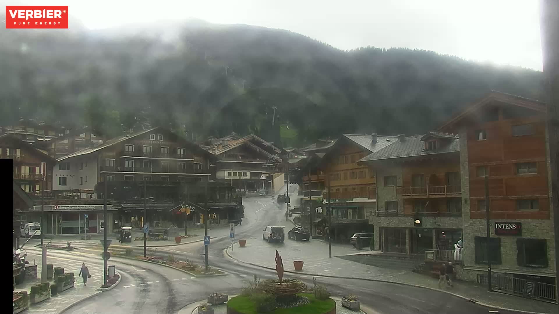 A village nestled in a mountain valley on a cloudy, wet day, with several buildings and cars visible on the wet streets.