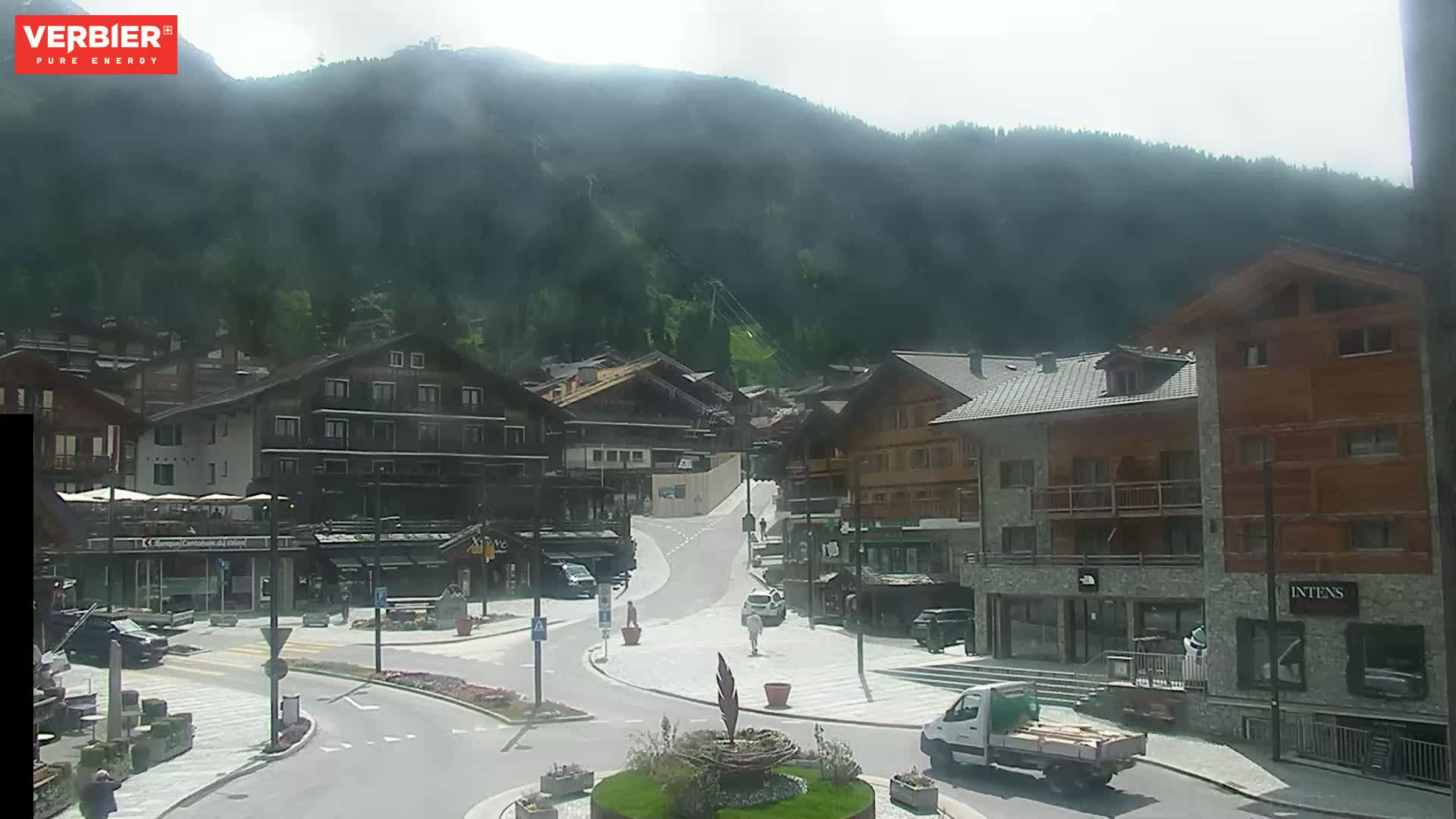 A hazy mountain village square with several buildings, a few cars, and pedestrians, is nestled at the base of a mountain under a hazy sky.