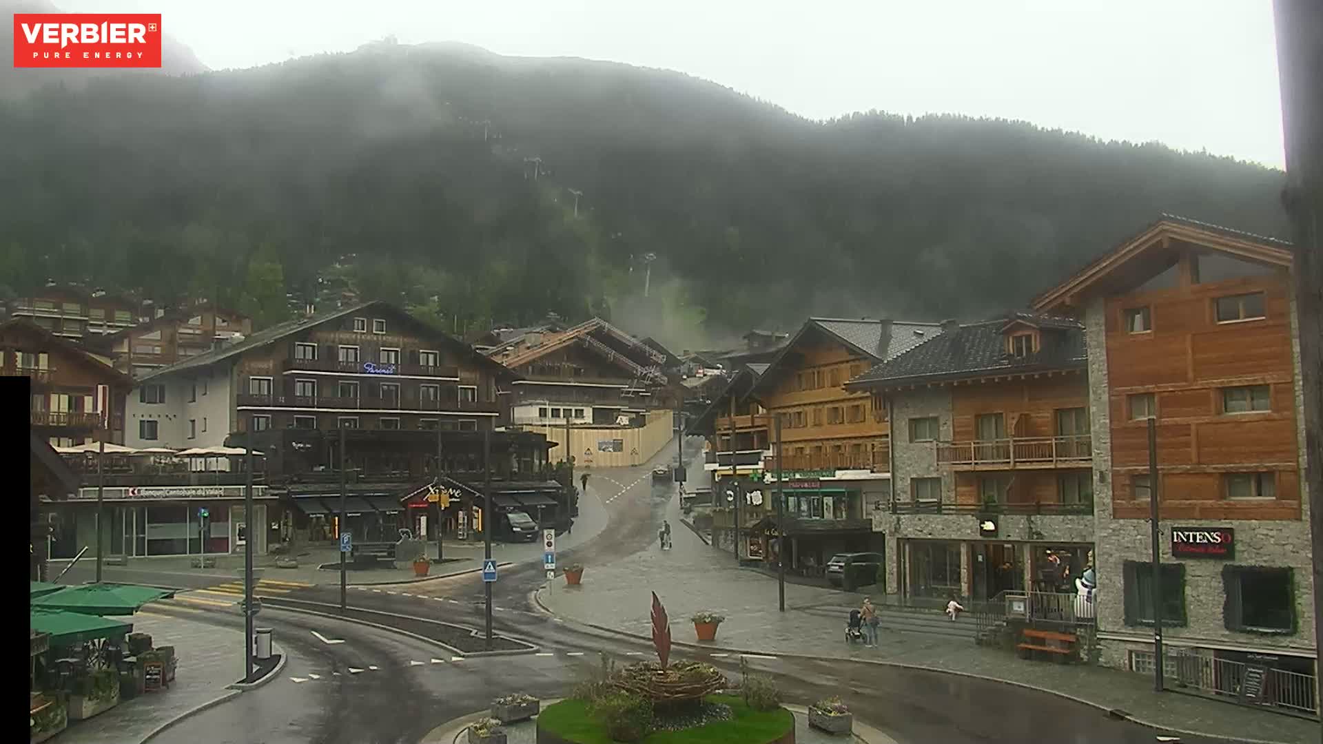 A rain-misted mountain village scene shows a town square with several multi-story buildings, wet streets, and a few pedestrians.