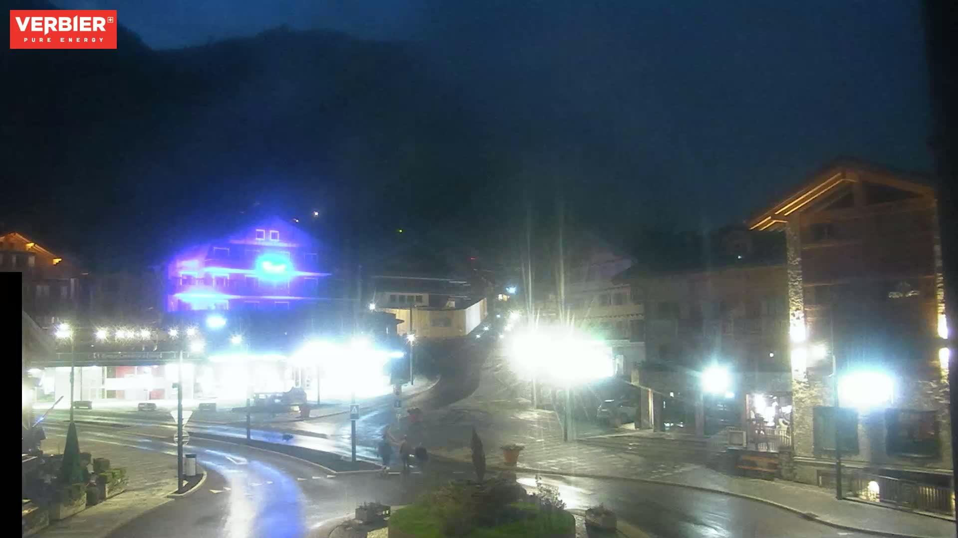 A nighttime view of a town square with wet streets, illuminated buildings, and streetlights under a dark sky.