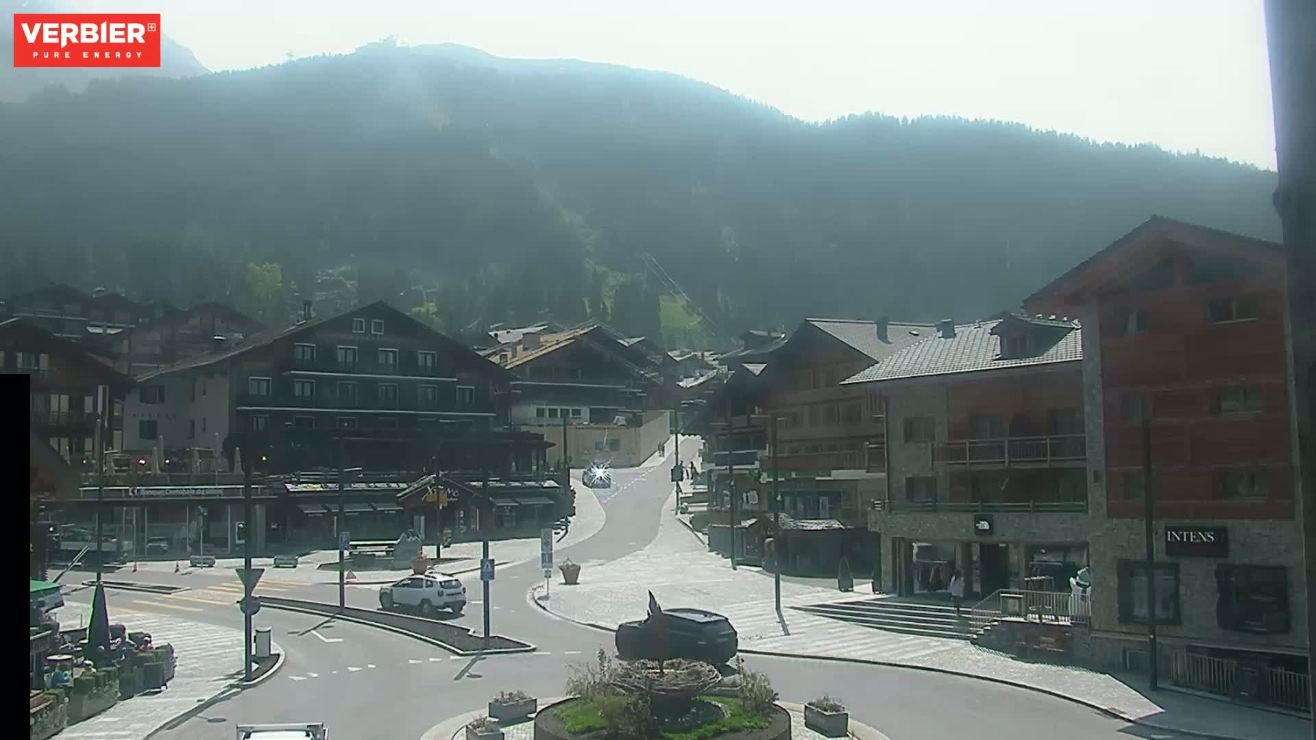 A hazy, sunny day reveals a mountain village square with several buildings, cars, and a small roundabout.