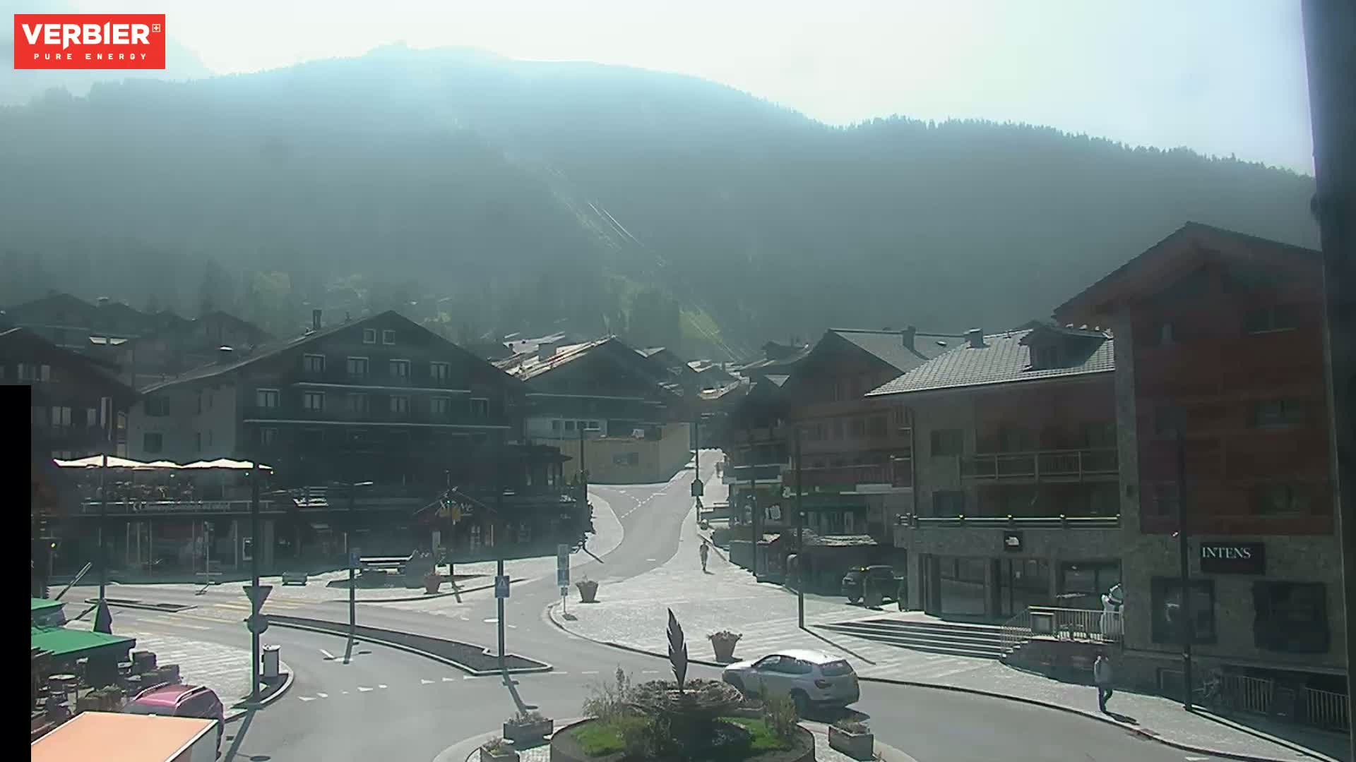 A hazy, sunny day reveals a mountain village with several buildings surrounding a paved intersection, a car parked nearby, and a few pedestrians walking.